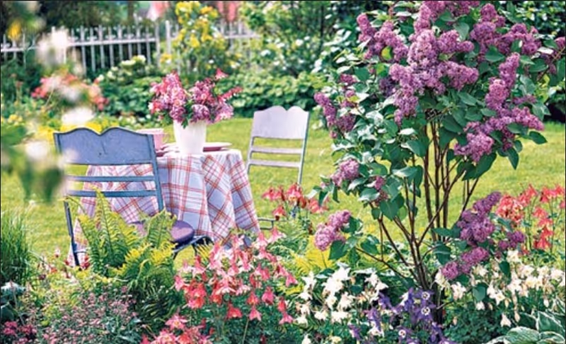A garden scene with a small table covered with a checkered tablecloth and a vase of purple flowers on top. Two chairs are placed around the table, surrounded by lush green grass and various colorful flowering plants including purple lilacs and pink and white flowers.
