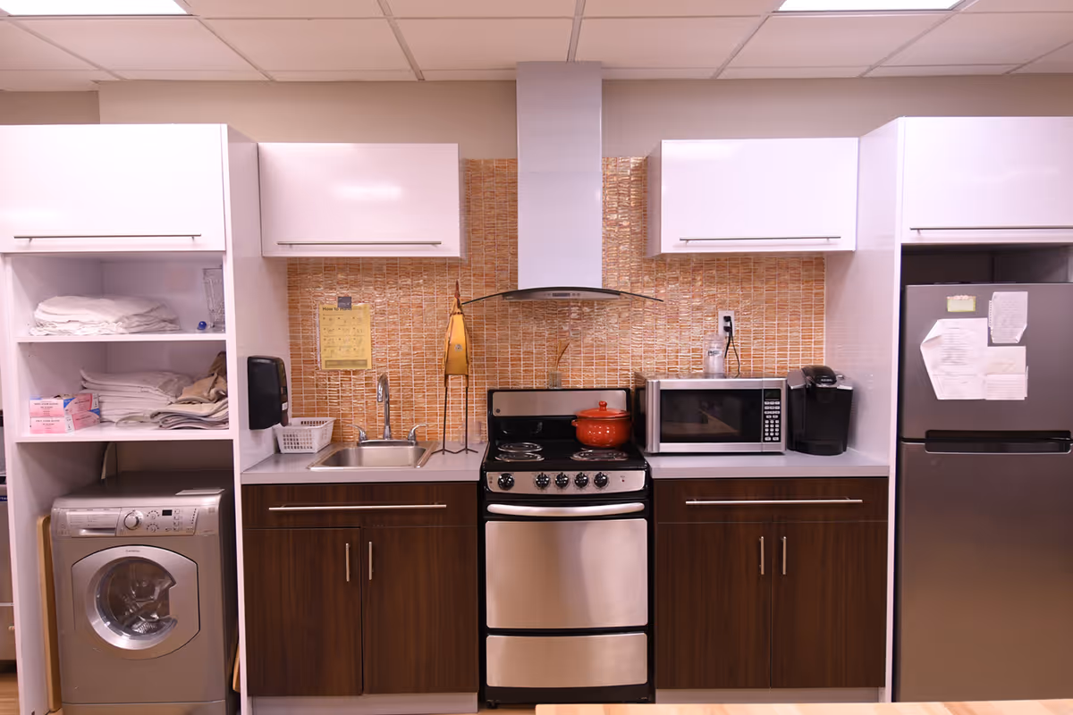 A kitchen area with a stainless steel stove, microwave, and refrigerator. There are wooden lower cabinets and white upper cabinets. To the left, there is a washing machine with shelves above holding folded towels and other items. The backsplash is made of small orange tiles, and there is a sink with a faucet in the middle.