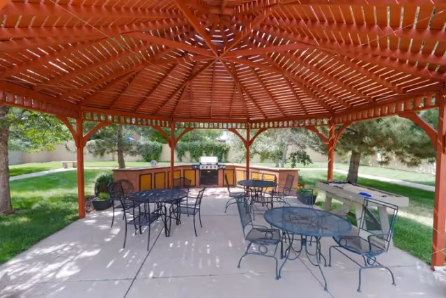Outdoor covered patio area with a red wooden pergola roof, several metal tables and chairs, a built-in grill, and a foosball table, surrounded by green grass and trees.