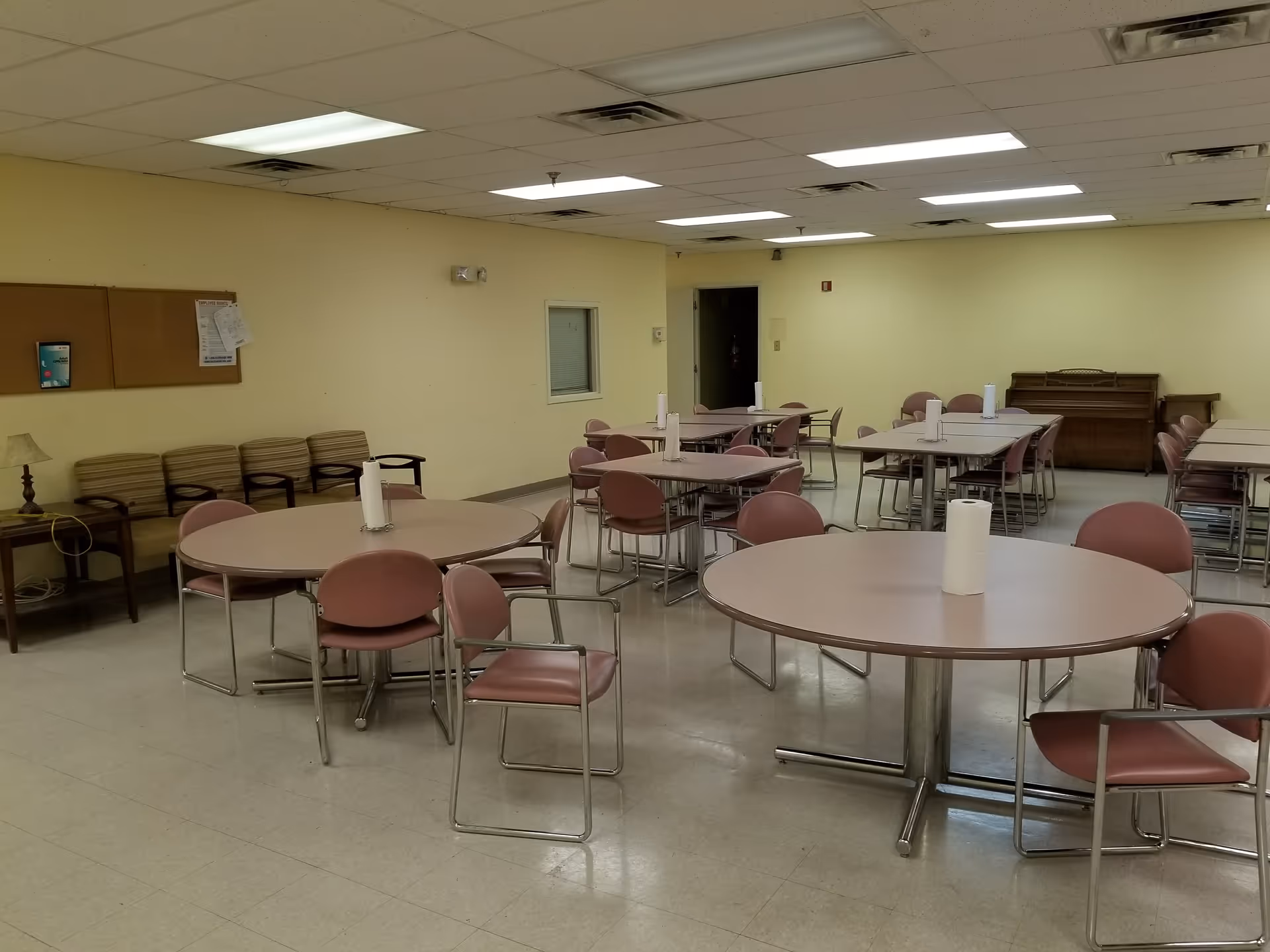 Empty dining room with round tables, pink chairs, and a piano in back.