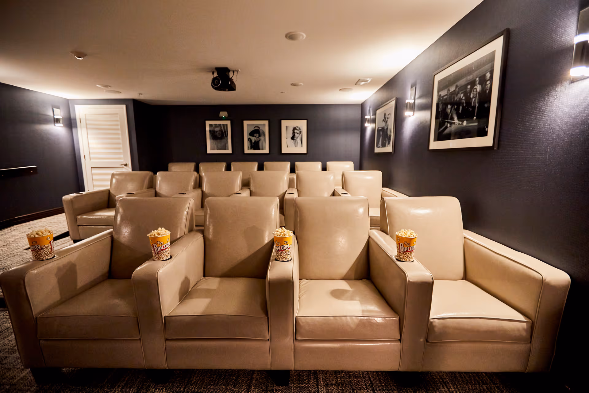 A small theater room with three rows of beige leather armchairs, each armchair having a cup of popcorn on the armrest. The walls are dark with framed black and white photographs, and there are wall-mounted lights providing soft illumination.
