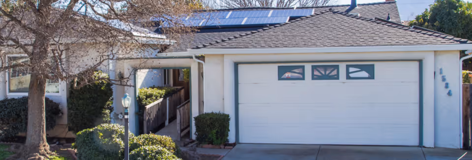 Front exterior of a single-story home with a two-car garage, driveway, entry ramp, and surrounding shrubs and trees.