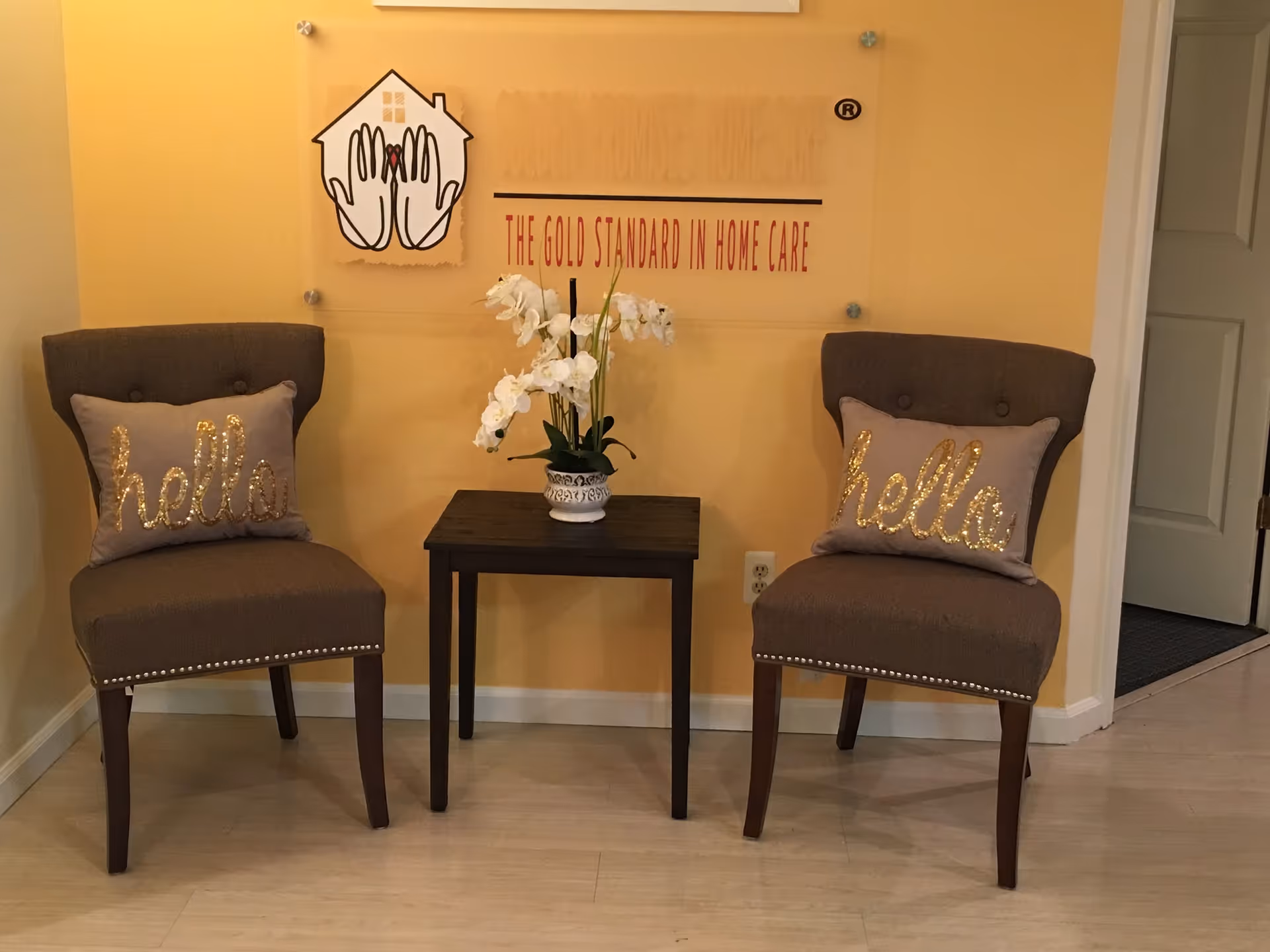 Two brown upholstered chairs flank a small table with white flowers in a yellow-walled lobby.