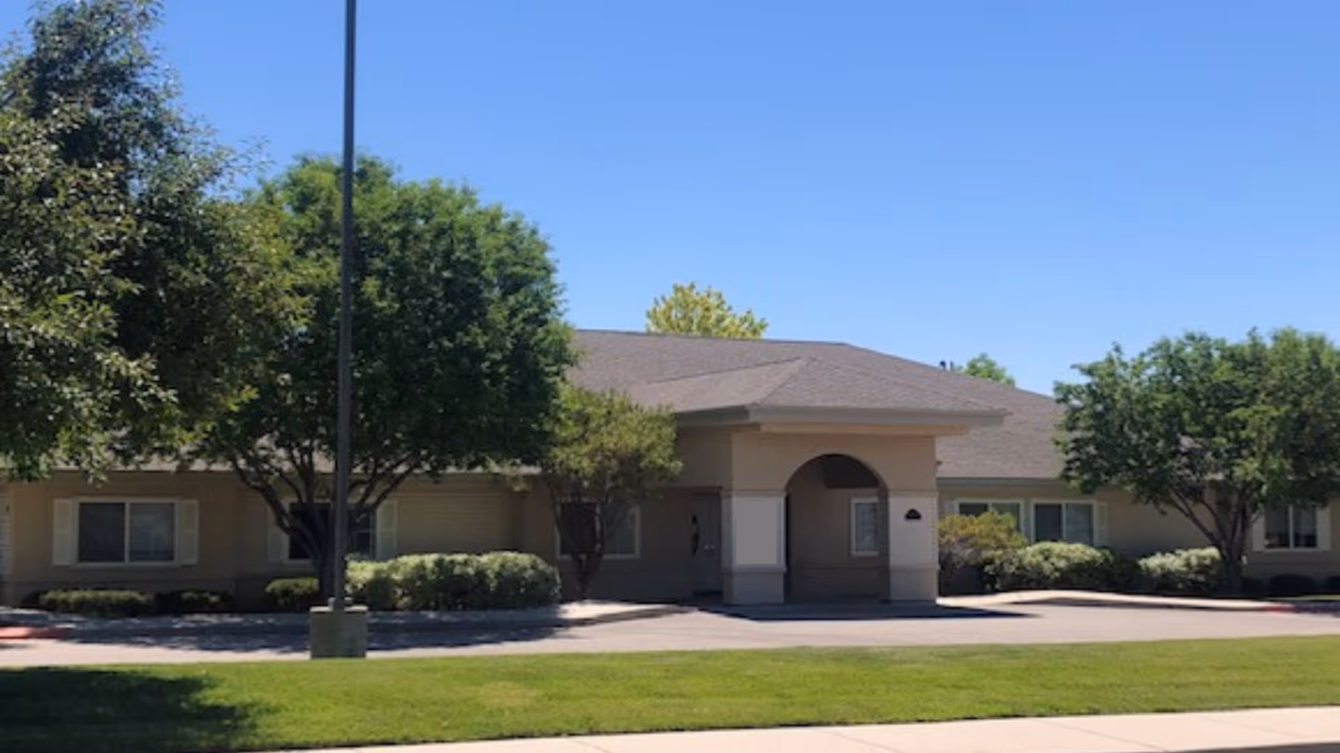 Single-story senior living facility exterior with an arched entrance, trees, and manicured lawn.