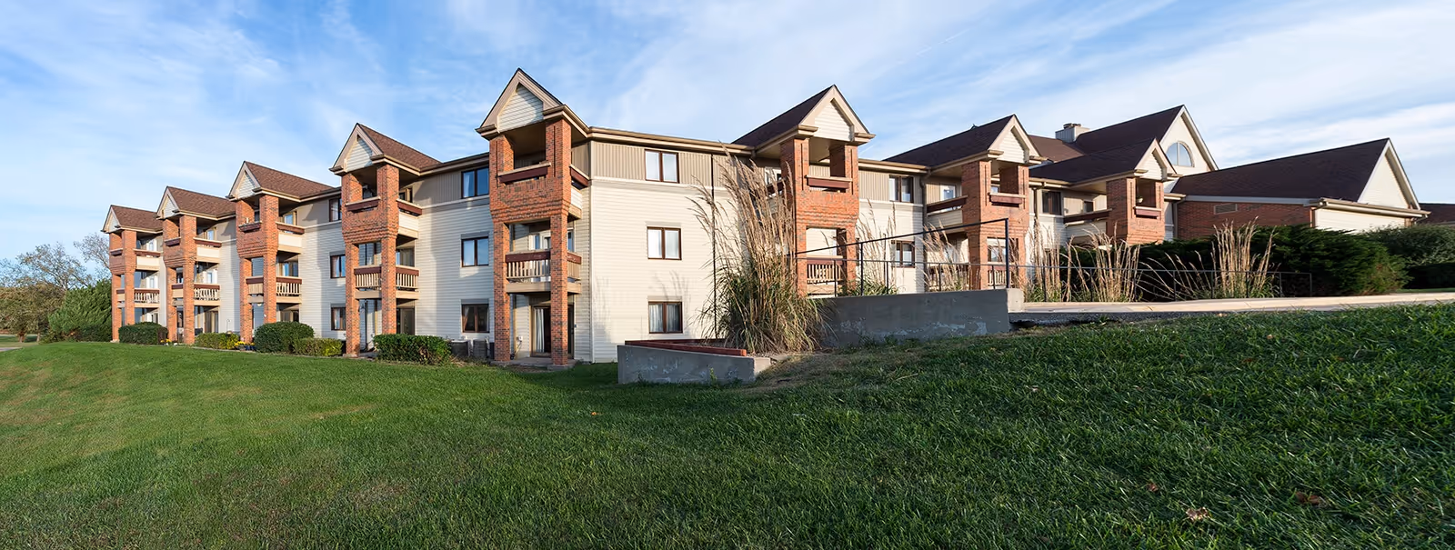 Exterior view of a multi-story senior living facility building with brick and beige siding, multiple balconies, and a well-maintained grassy lawn in the foreground under a clear blue sky.