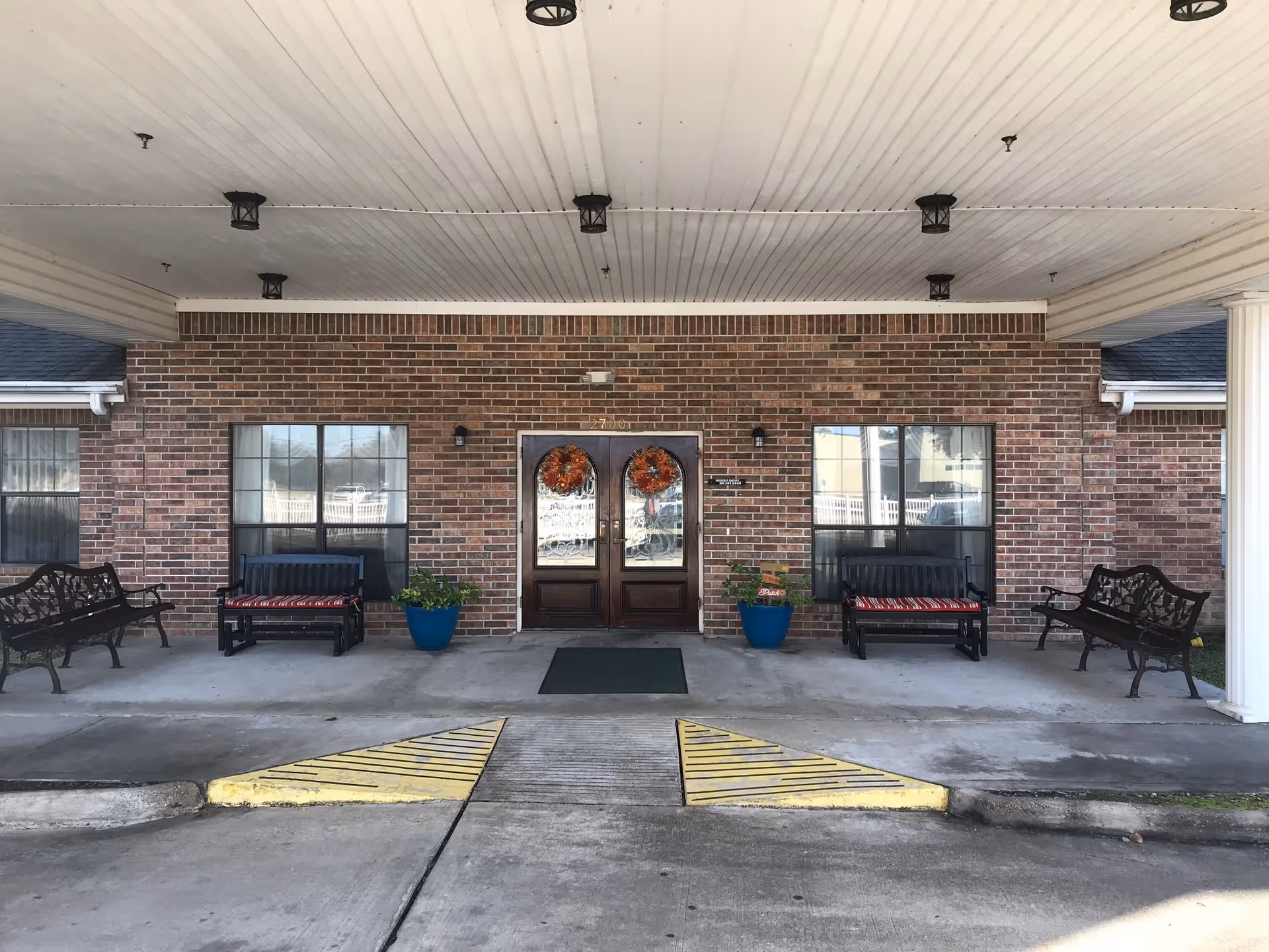 Entrance of a brick building with double wooden doors decorated with autumn wreaths. There are two windows on either side of the doors, each with a bench and a blue planter with green plants. The area is covered by a white ceiling with multiple light fixtures, and there is a concrete driveway with yellow painted ramps leading to the entrance.