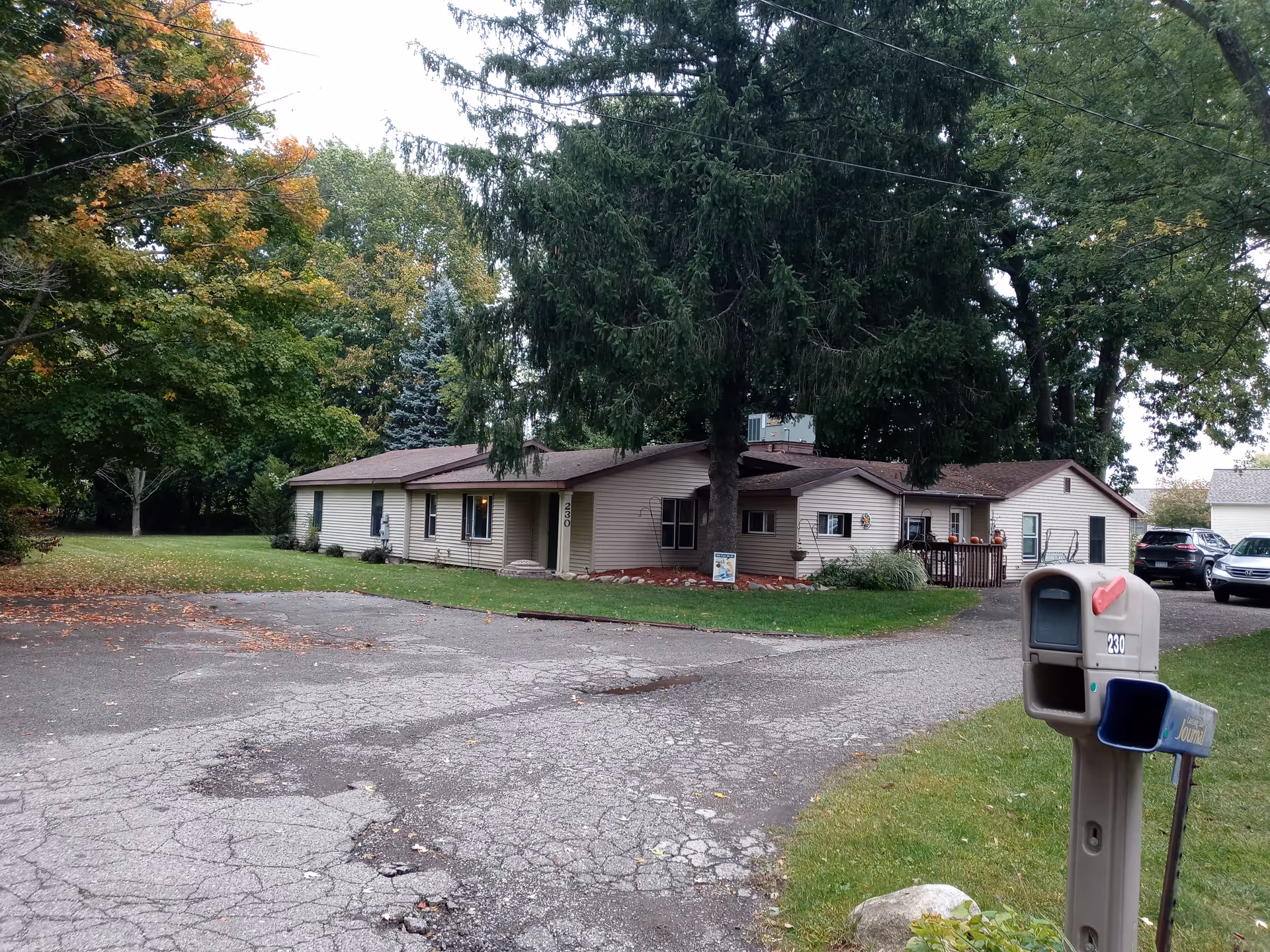 One-story beige care facility with a driveway, mailbox, and tall trees in front yard.