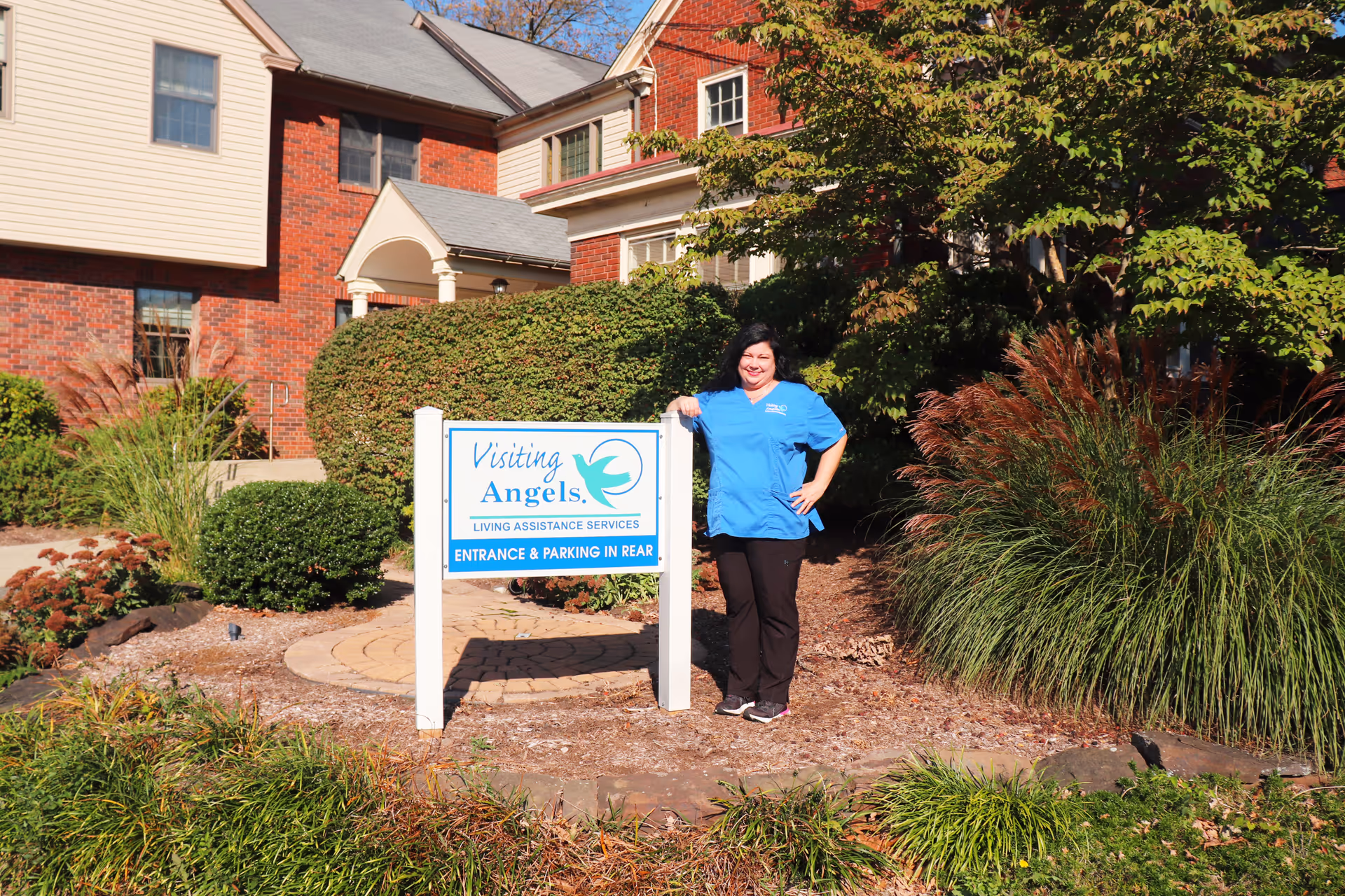 Caregiver in blue scrubs stands beside Visiting Angels sign at entrance.