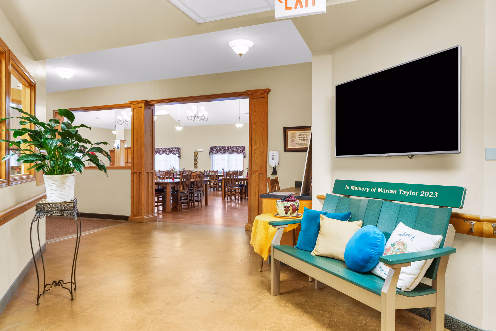 Interior lobby area with teal bench, potted plant, large TV, and doorway to dining room.
