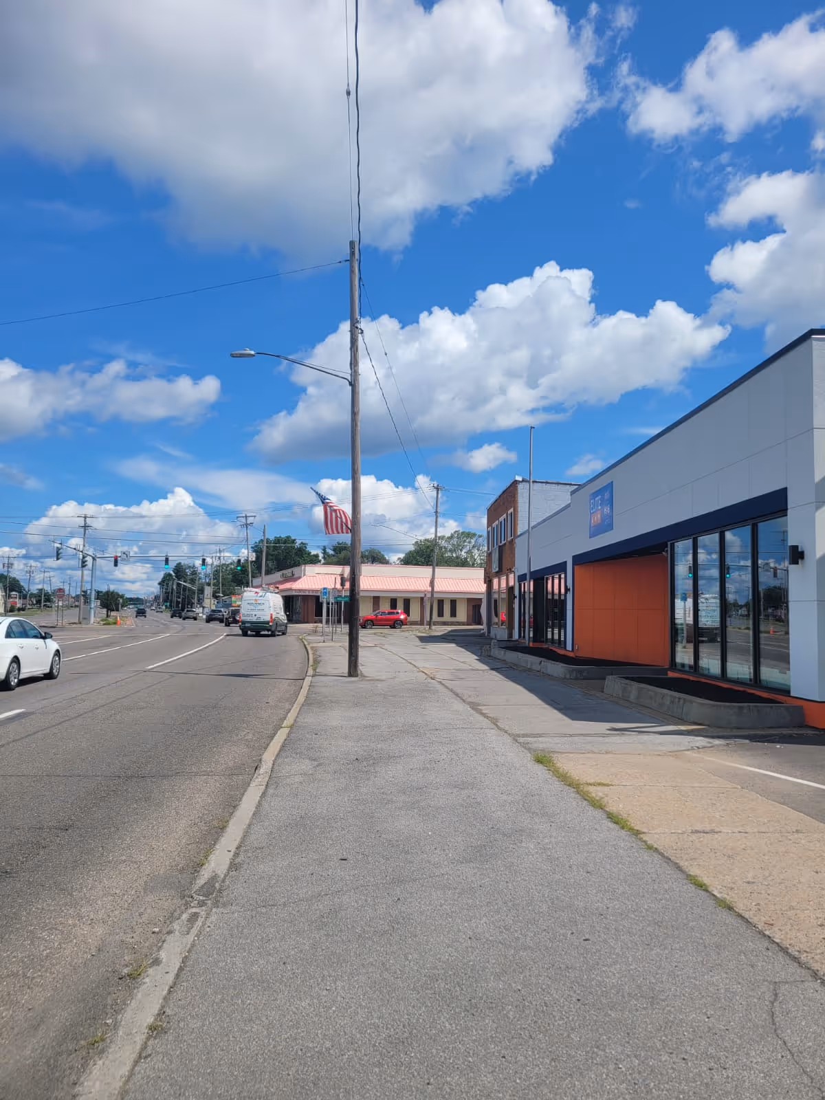 Wide street view with sidewalk, utility pole, and storefronts under a bright blue sky.