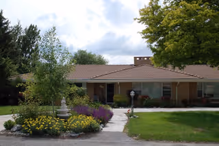 Single-story residential building with a brown roof surrounded by green trees and a garden with yellow and purple flowers in front. A concrete pathway leads to the entrance of the building under a cloudy sky.