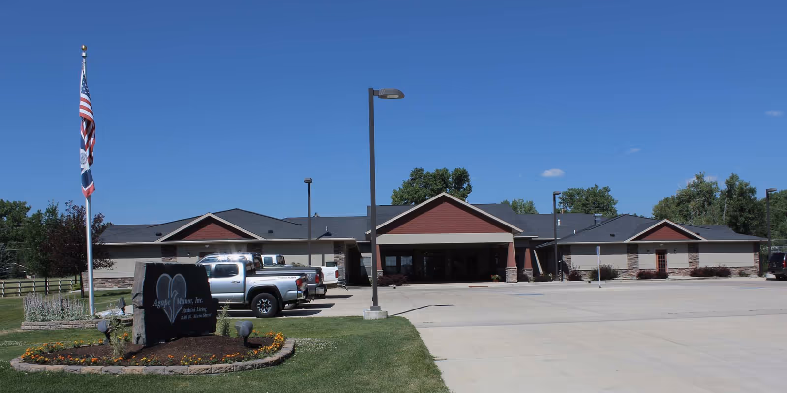 Front exterior of a single-story senior living facility with a driveway, parked trucks, a flagpole, and a stone sign reading Agape Manor, Inc.