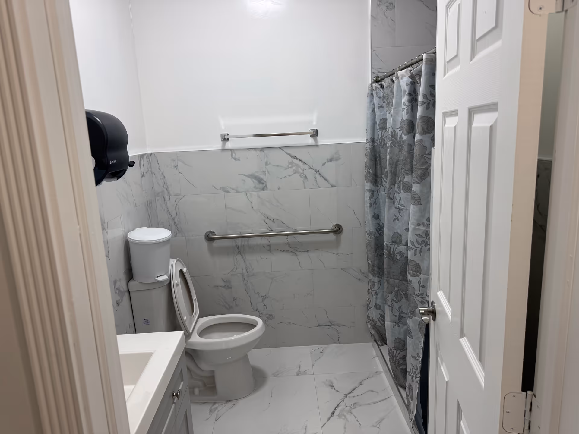 Small marble-tiled bathroom with toilet, sink, grab bars, and shower curtain.