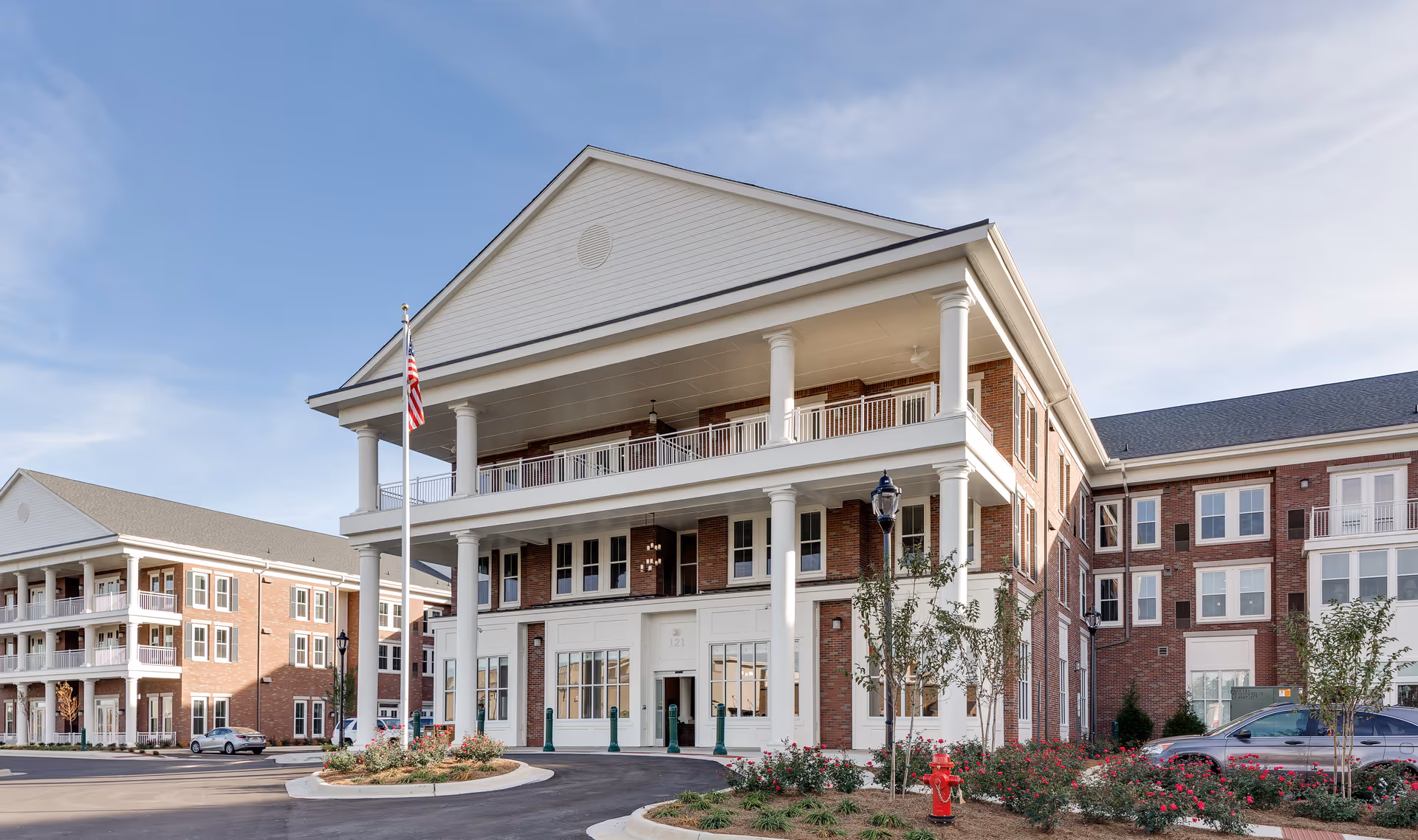 Front exterior of a multi-story brick senior living building with white columns, covered balconies, landscaping, and an American flag at the entrance.
