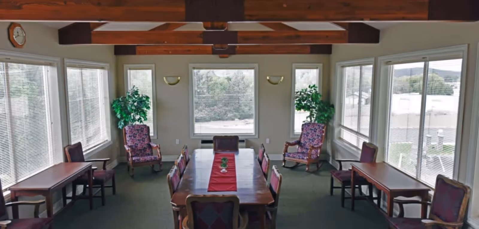 Sunlit dining room with a long wooden table, chairs, and potted plants by large windows.