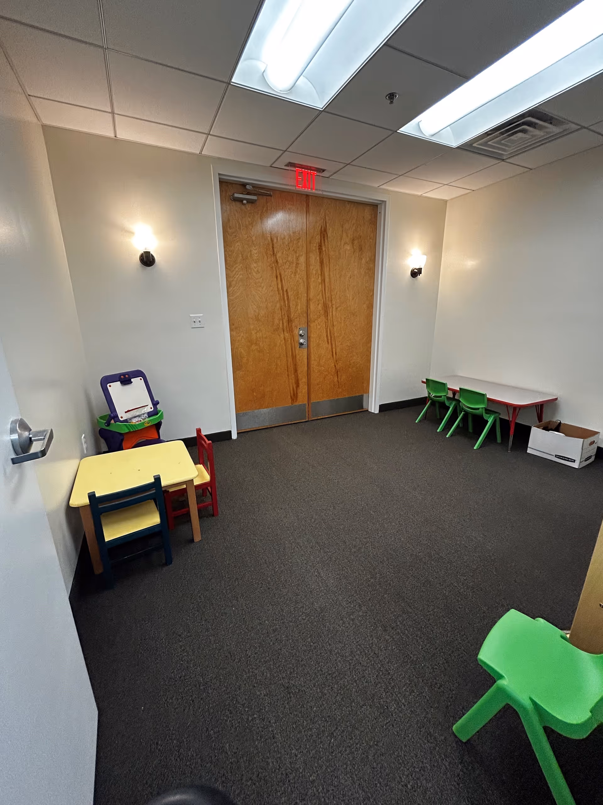 Small activity room with colorful kid-sized tables and chairs, back door and bright lights.