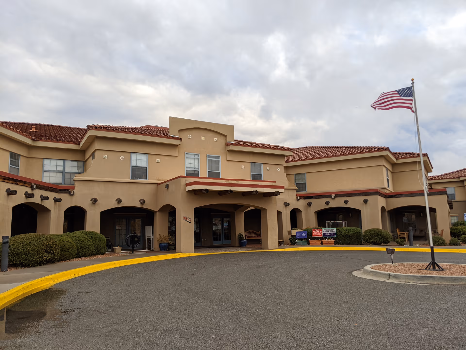 Beige senior living facility exterior with arches, red-tiled roof, and flagpole.