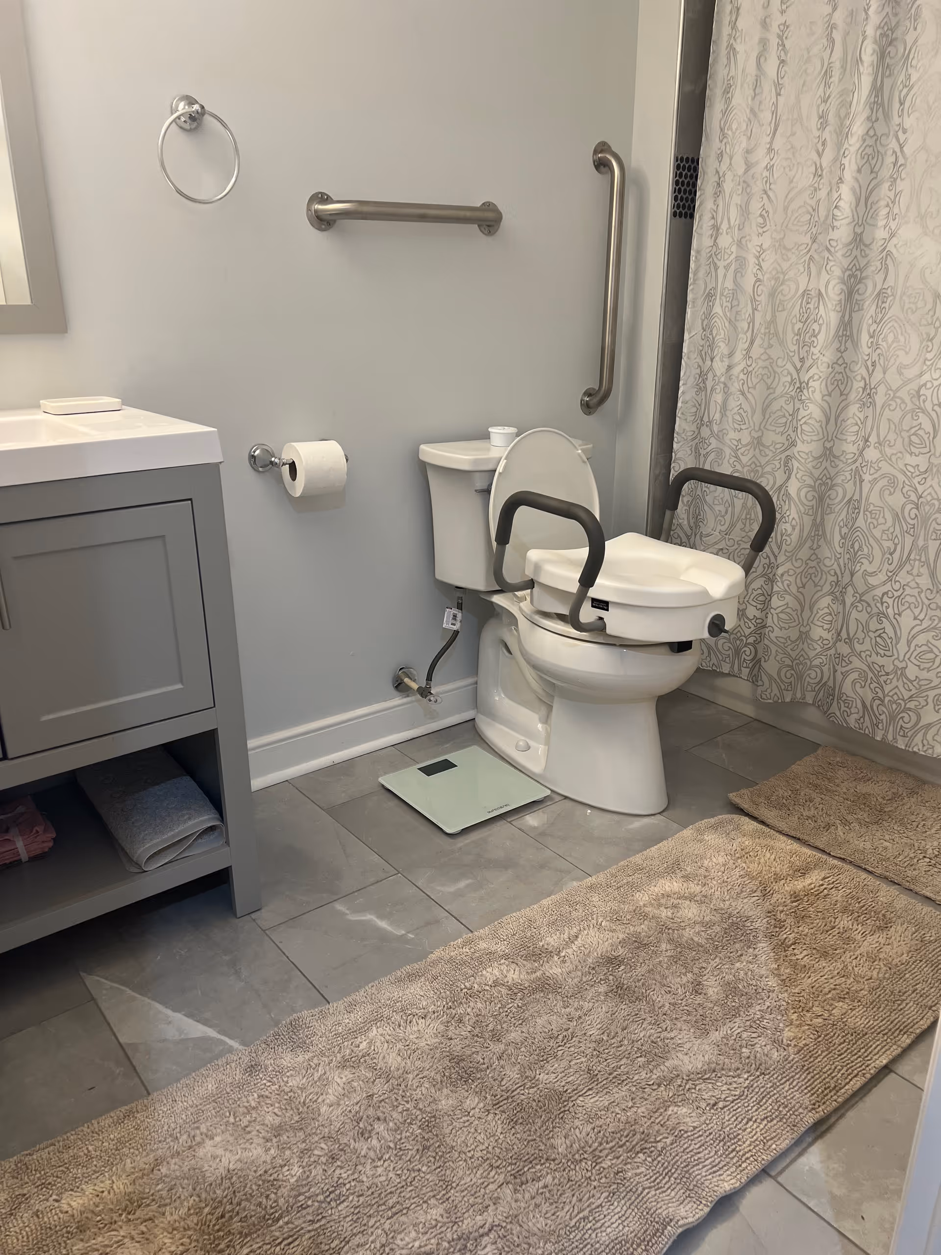Accessible bathroom with toilet, grab bars, sink, and shower curtain in a gray facility.
