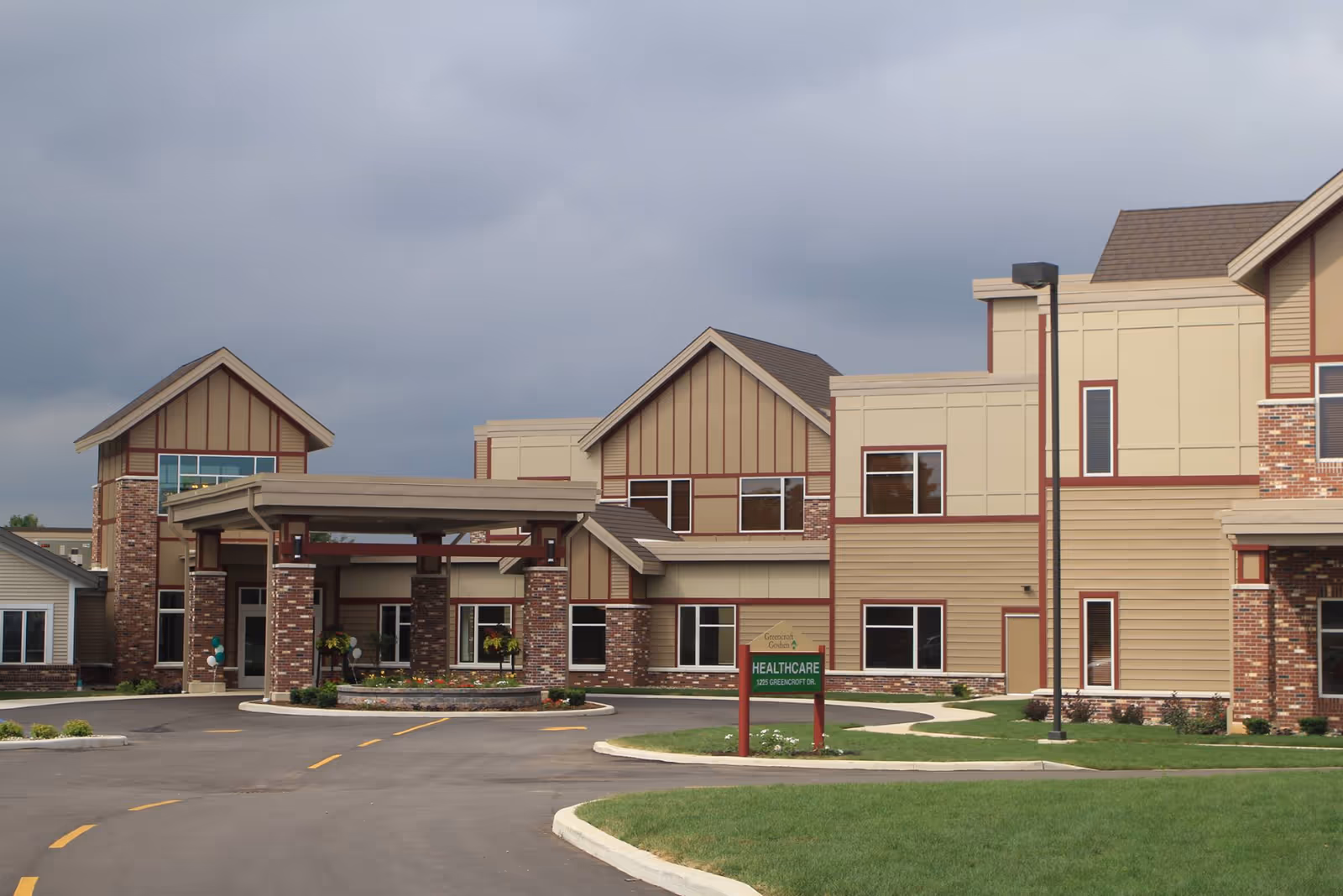 Exterior view of Greencroft Goshen healthcare facility showing a large building with beige and brick facade, multiple windows, and a covered entrance with pillars. There is a green sign in front that reads 'Greencroft Goshen HEALTHCARE 1225 GREENCROFT DR.' The sky is cloudy and the driveway curves toward the entrance.