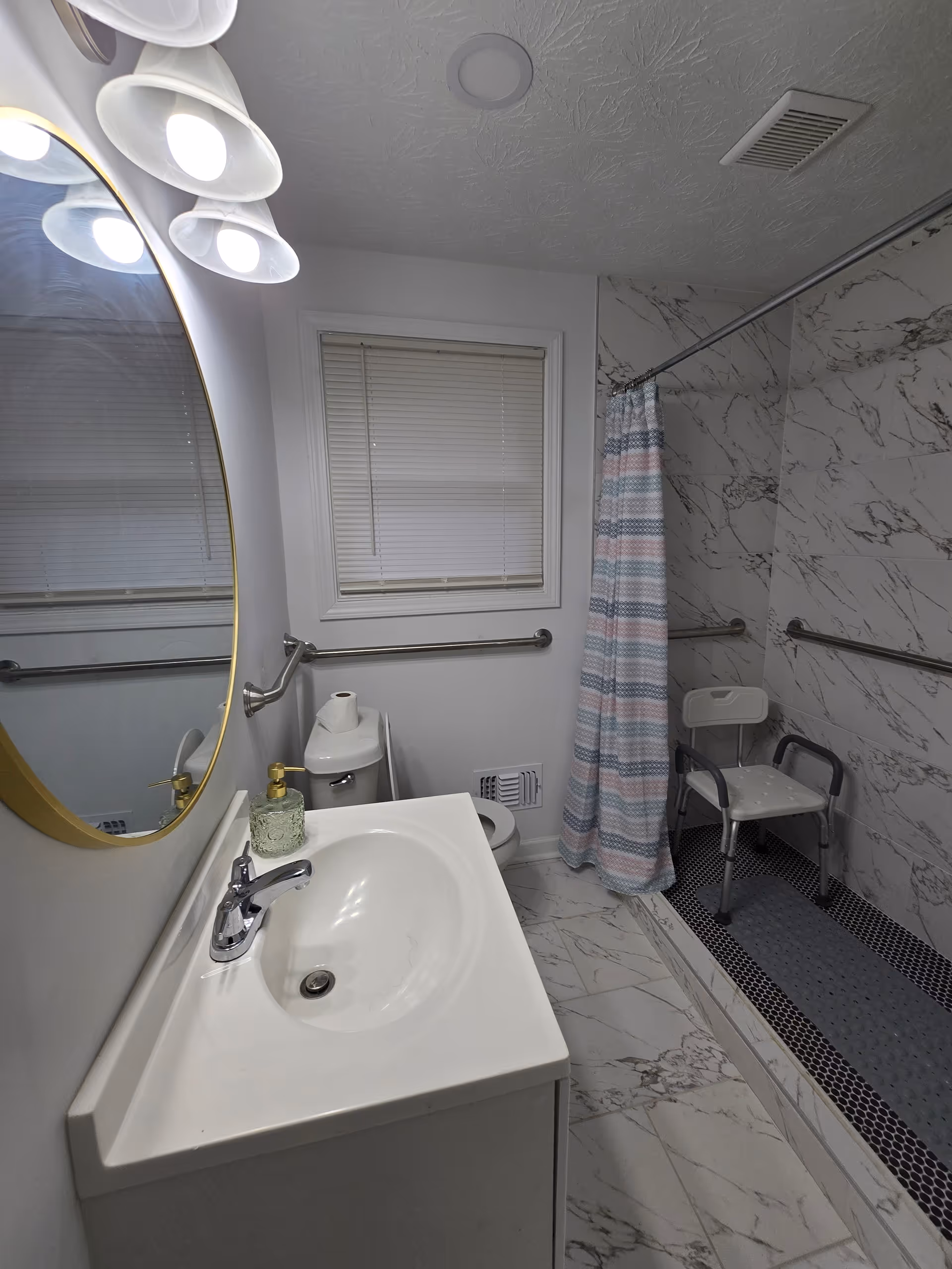Bathroom with sink, mirror, window, shower chair, and wall-mounted grab bars.