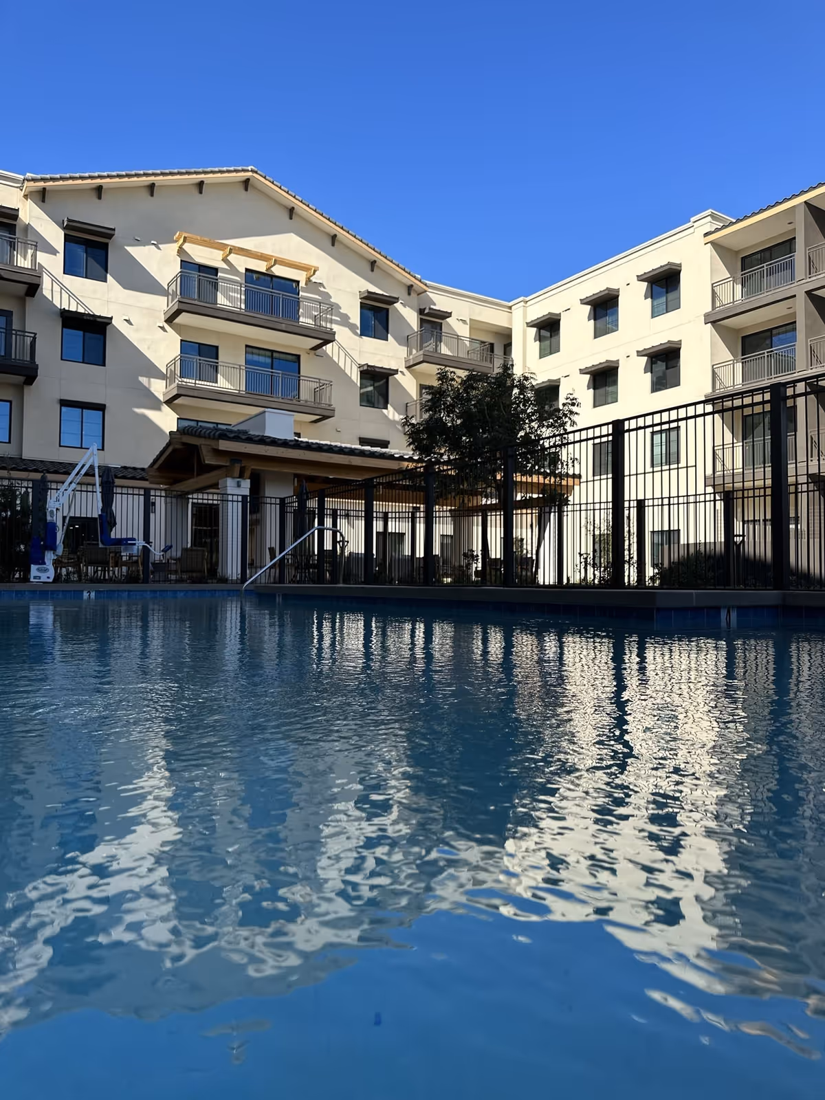Outdoor swimming pool with water reflections in the foreground and a multi-story senior living building with balconies under a clear blue sky.