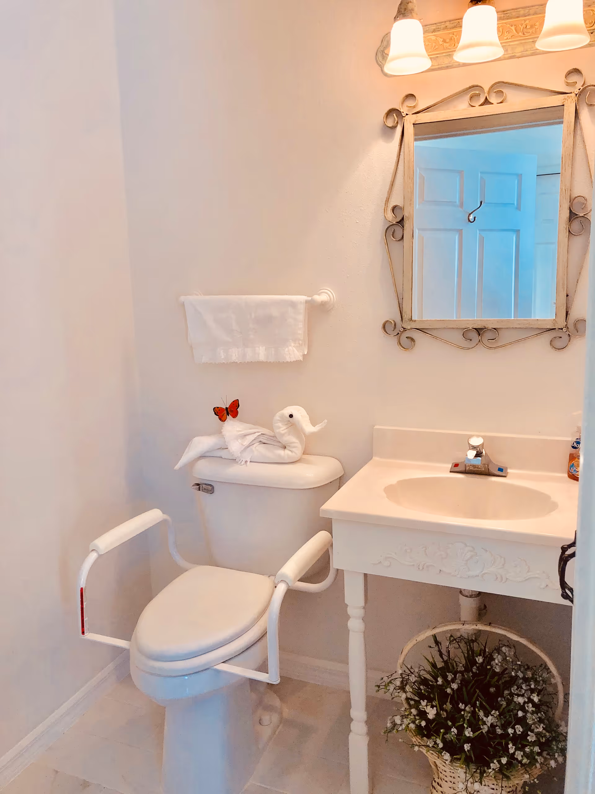Small decorated bathroom with a toilet fitted with safety rails and a towel-swan on the tank, a sink vanity with mirror, and a floral basket beneath.