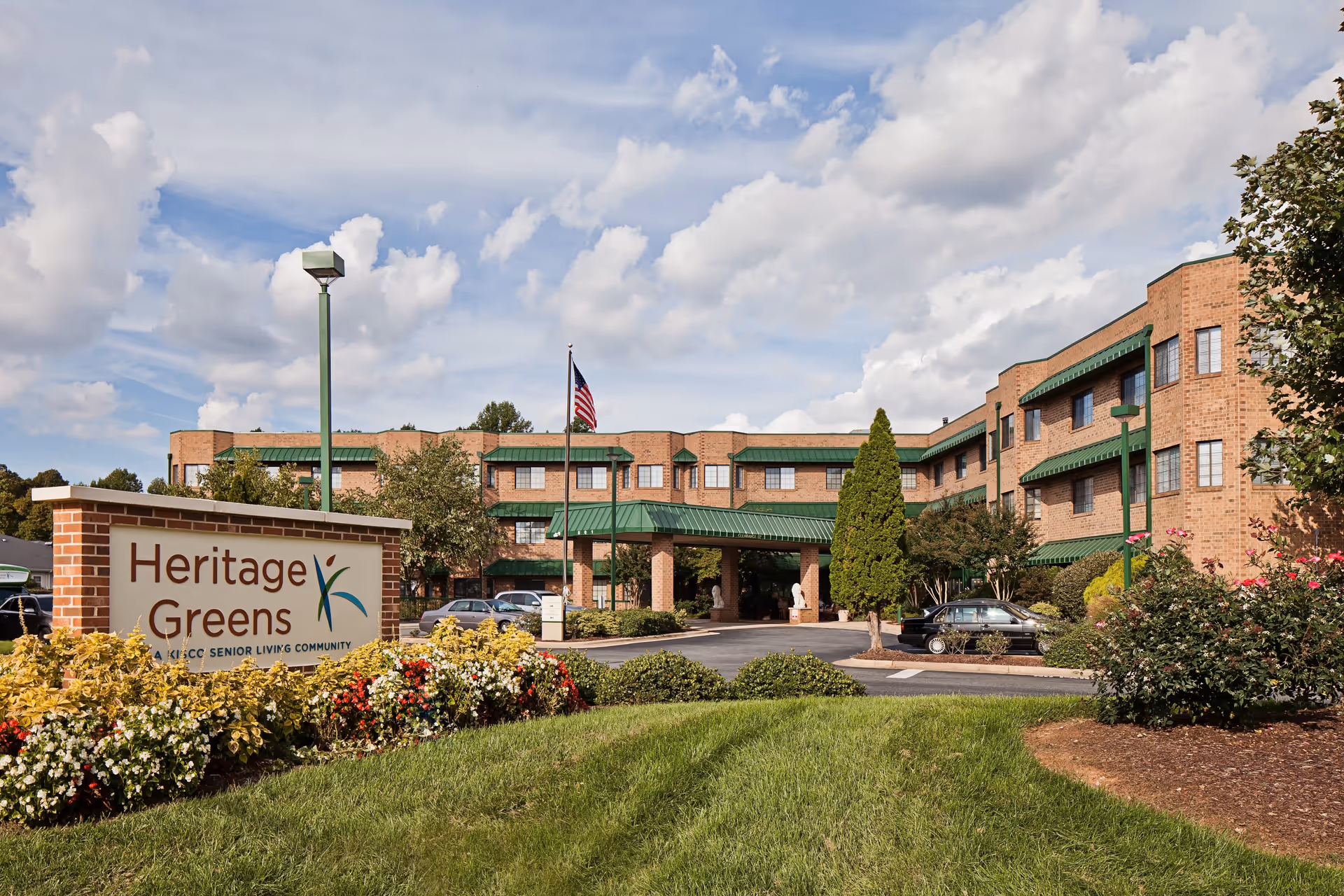 Exterior view of Heritage Greens senior living community building with a brick facade, green awnings, and a covered entrance. There is a sign in the foreground with the facility name surrounded by colorful flowers and greenery. Several cars are parked near the entrance under a partly cloudy sky.