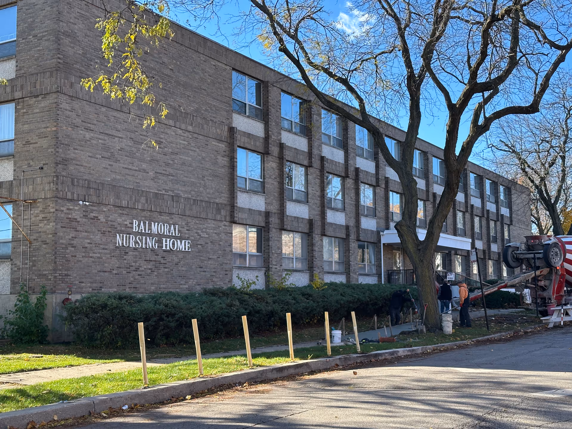 Exterior view of Balmoral Nursing Home, a three-story brick building with multiple windows. There are trees with bare branches and some workers near the entrance, along with construction equipment on the right side of the image. The sky is clear and blue.