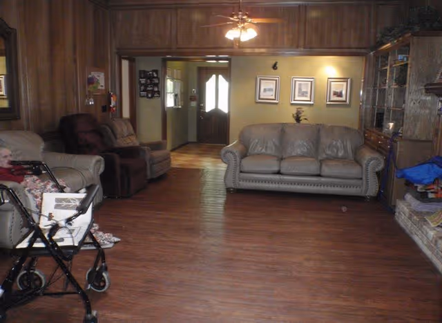 Cozy living room in a senior facility with leather sofa, recliners, wood paneling, and a walker.
