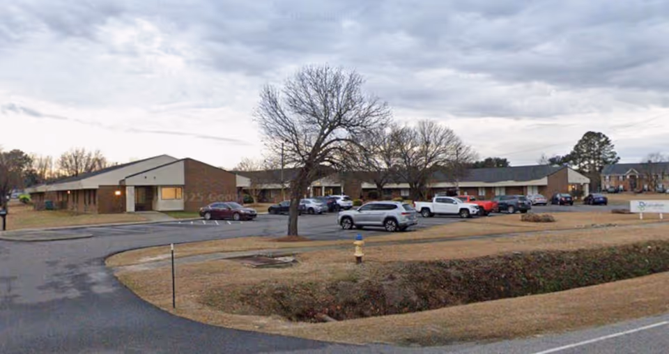 Single-story brick nursing and rehabilitation center exterior with parked cars and leafless trees.