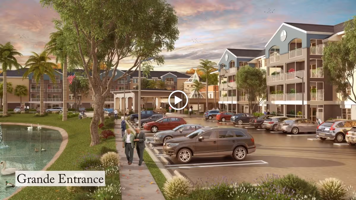 View of the exterior of a senior living facility named Discovery Village At Boynton Beach during sunset. The image shows a large parking lot with several parked cars, a landscaped pond with ducks and swans on the left, palm trees, and a wide sidewalk where two elderly people are walking. The multi-story building has balconies and a grand entrance with a covered drop-off area. The sky is partly cloudy with warm colors.