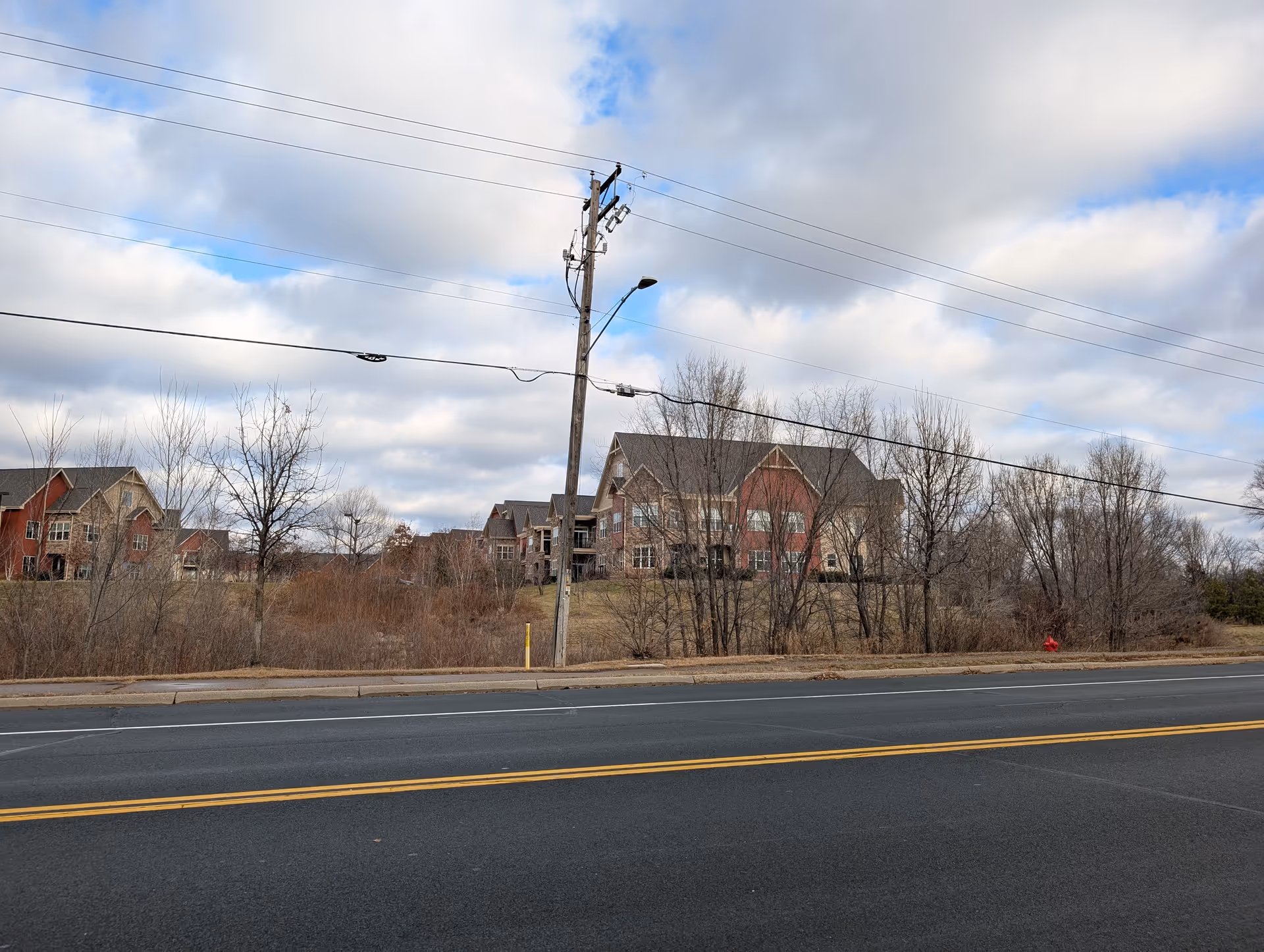 View of a senior living facility named Founders Ridge from across a road. The facility consists of multiple multi-story buildings with red and beige exteriors, surrounded by leafless trees and dry grass under a cloudy sky.