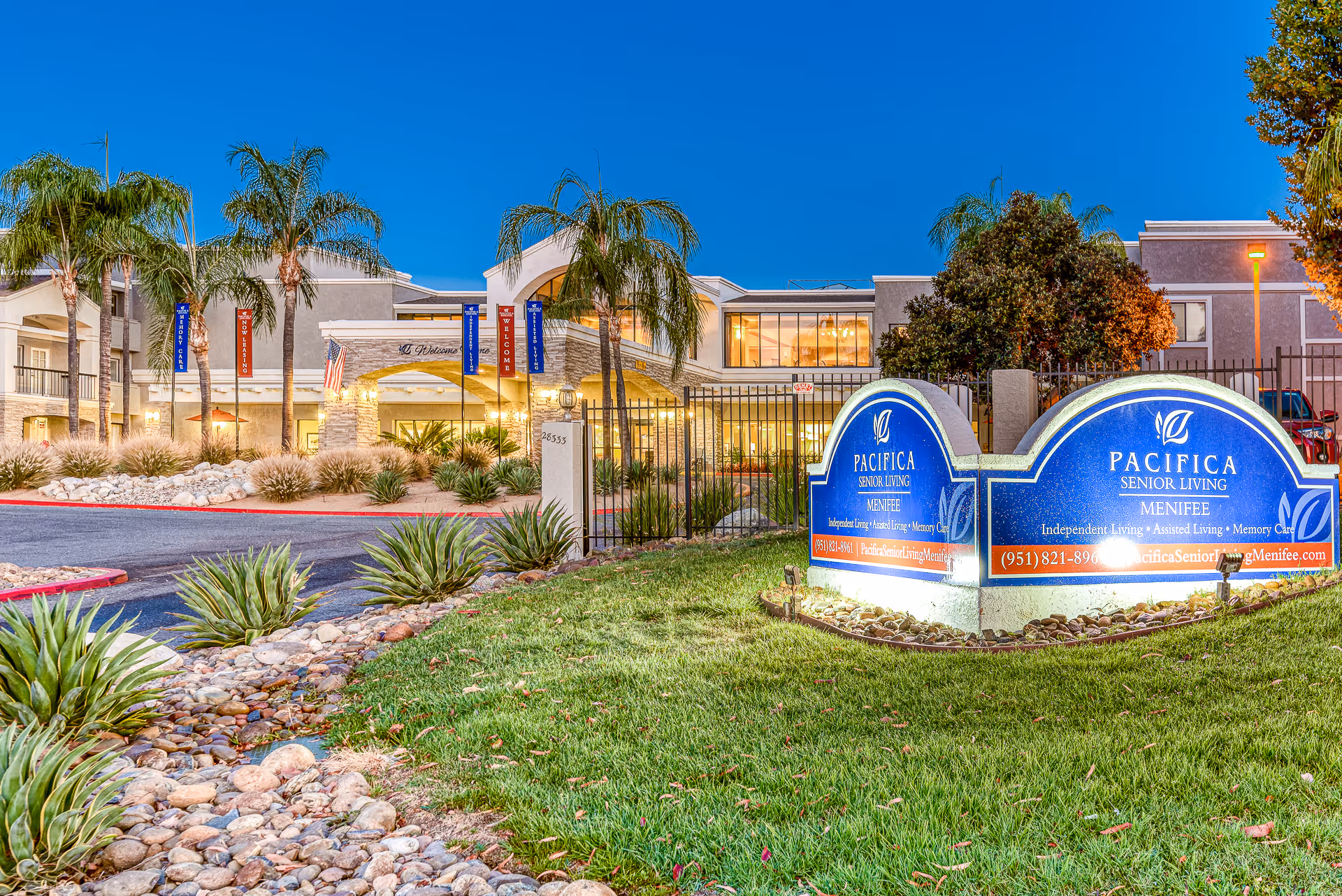 Front exterior of Pacifica Senior Living Menifee with illuminated entrance signs, palm trees, and landscaped driveway at dusk.