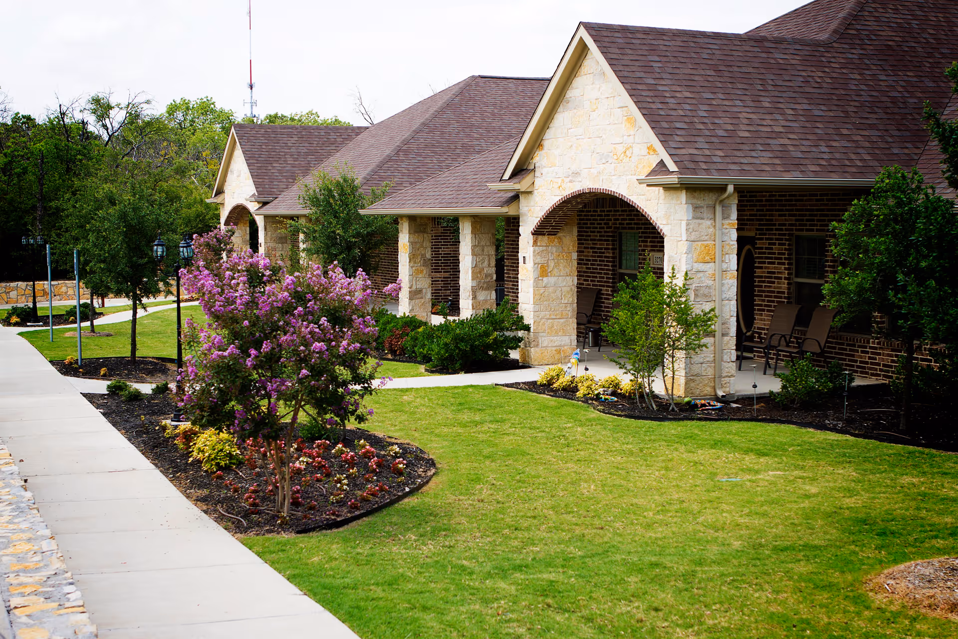 Exterior view of a single-story assisted living facility building with stone and brick facade, a well-maintained lawn, landscaped flower beds with blooming purple flowers, and a concrete sidewalk leading alongside the building.
