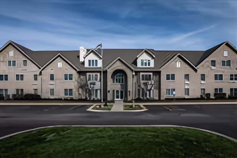 Front exterior view of a large, multi-story building with multiple windows and a central entrance under a peaked roof, set against a clear blue sky with a paved driveway and green lawn in front.