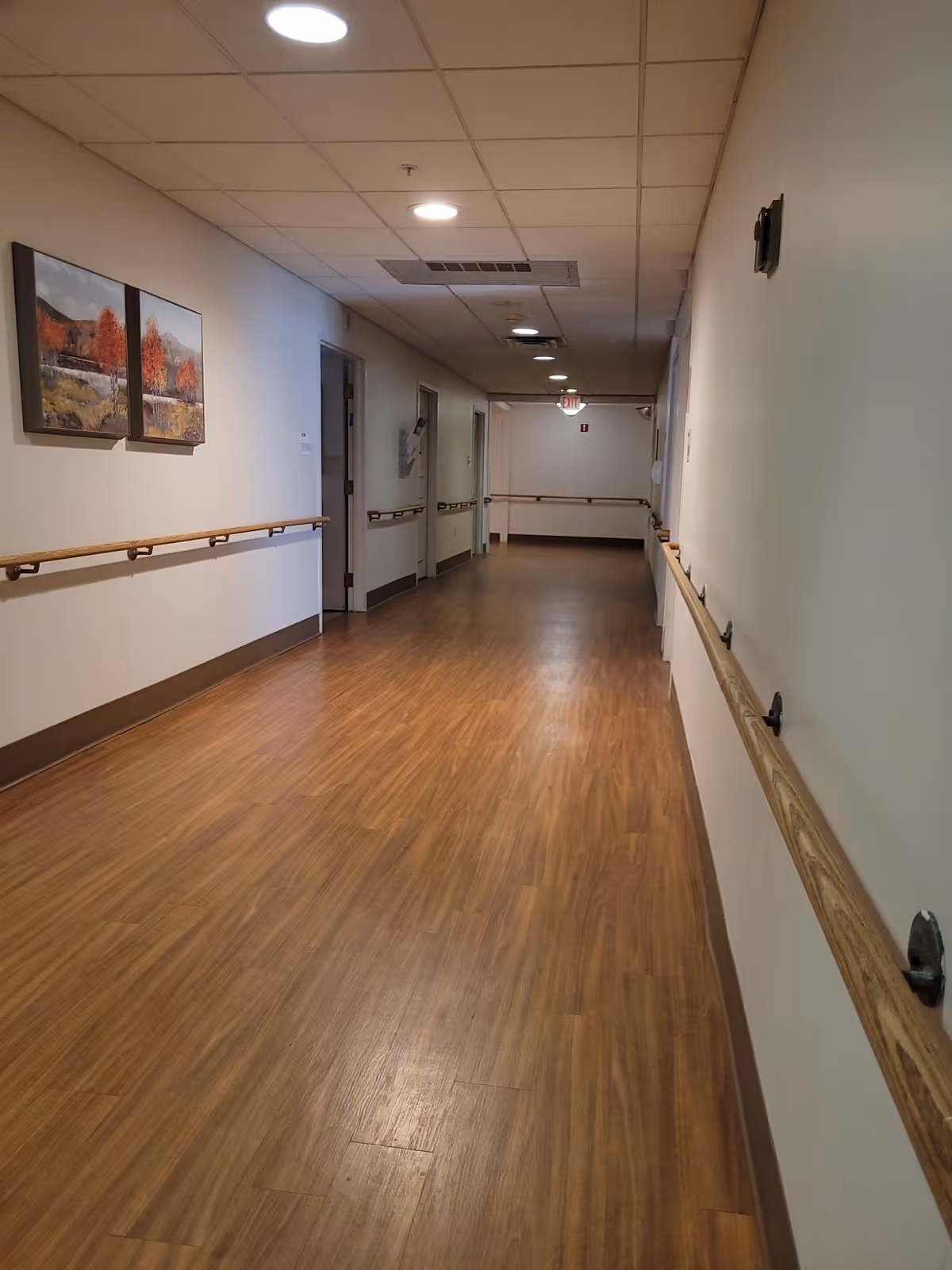 Long, well-lit hallway in a senior living facility with wood floors and handrails.