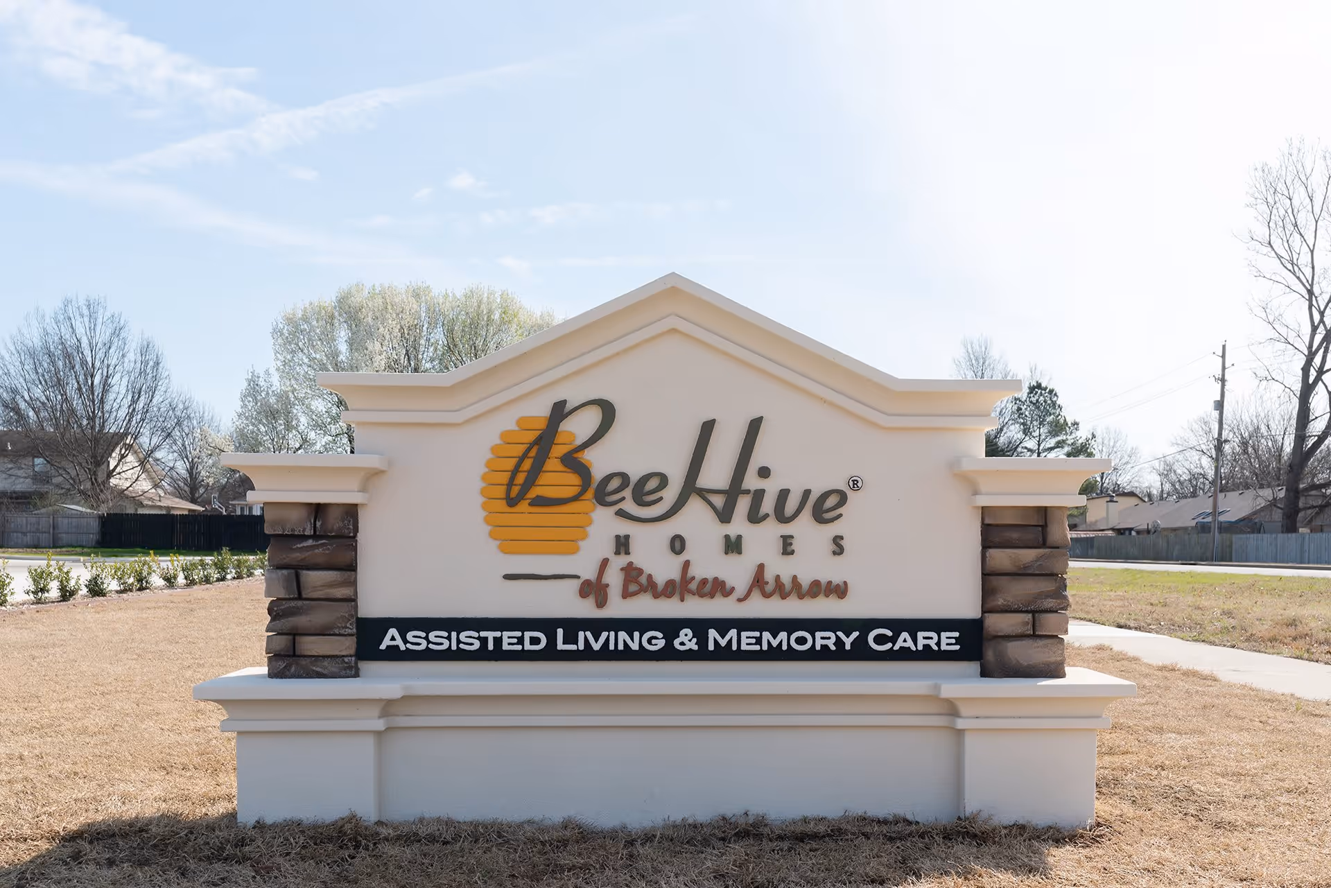 Outdoor sign for BeeHive Homes of Broken Arrow, an assisted living and memory care facility, with a clear sky and trees in the background.