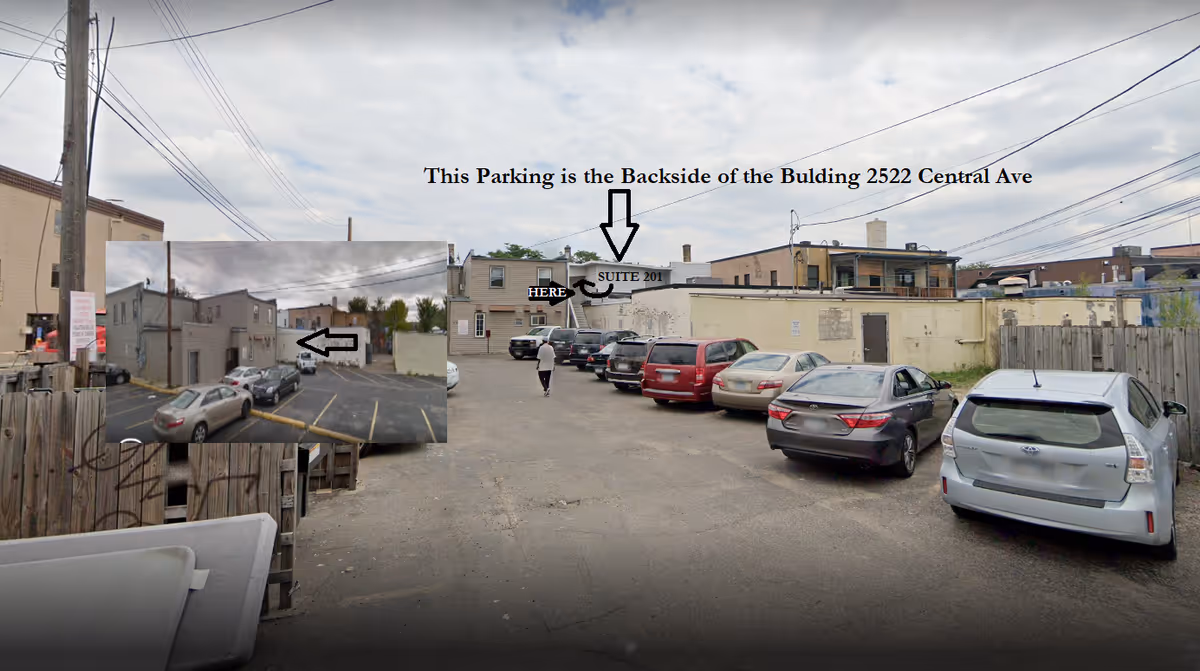 Rear parking lot with several cars and a person walking toward a beige building.