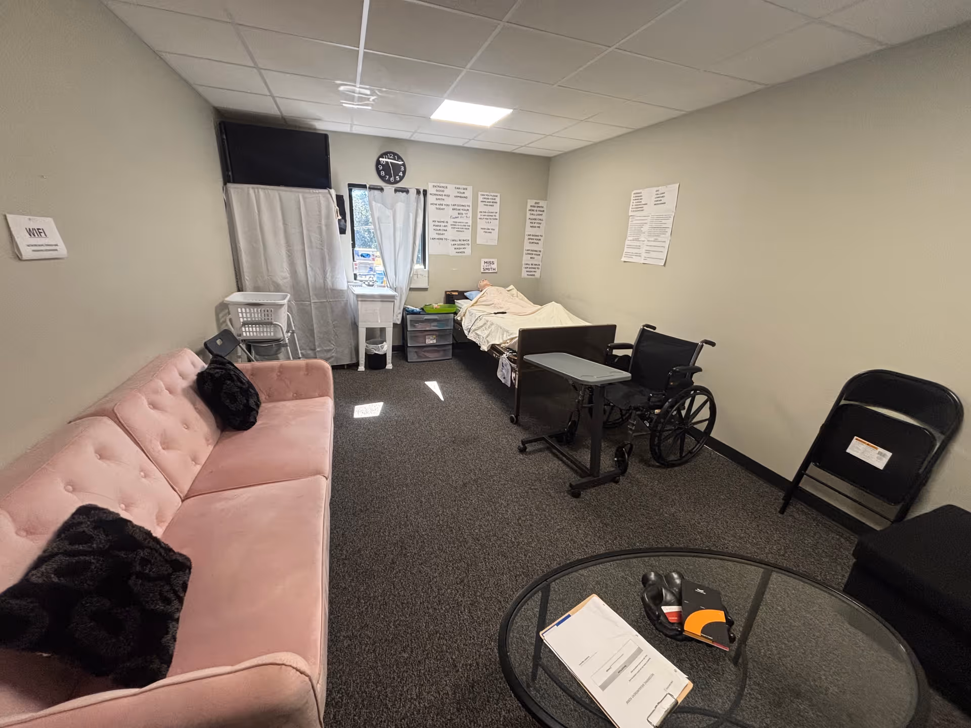 A small hospital-like living area with a pink sofa, bed, wheelchair, and tables.