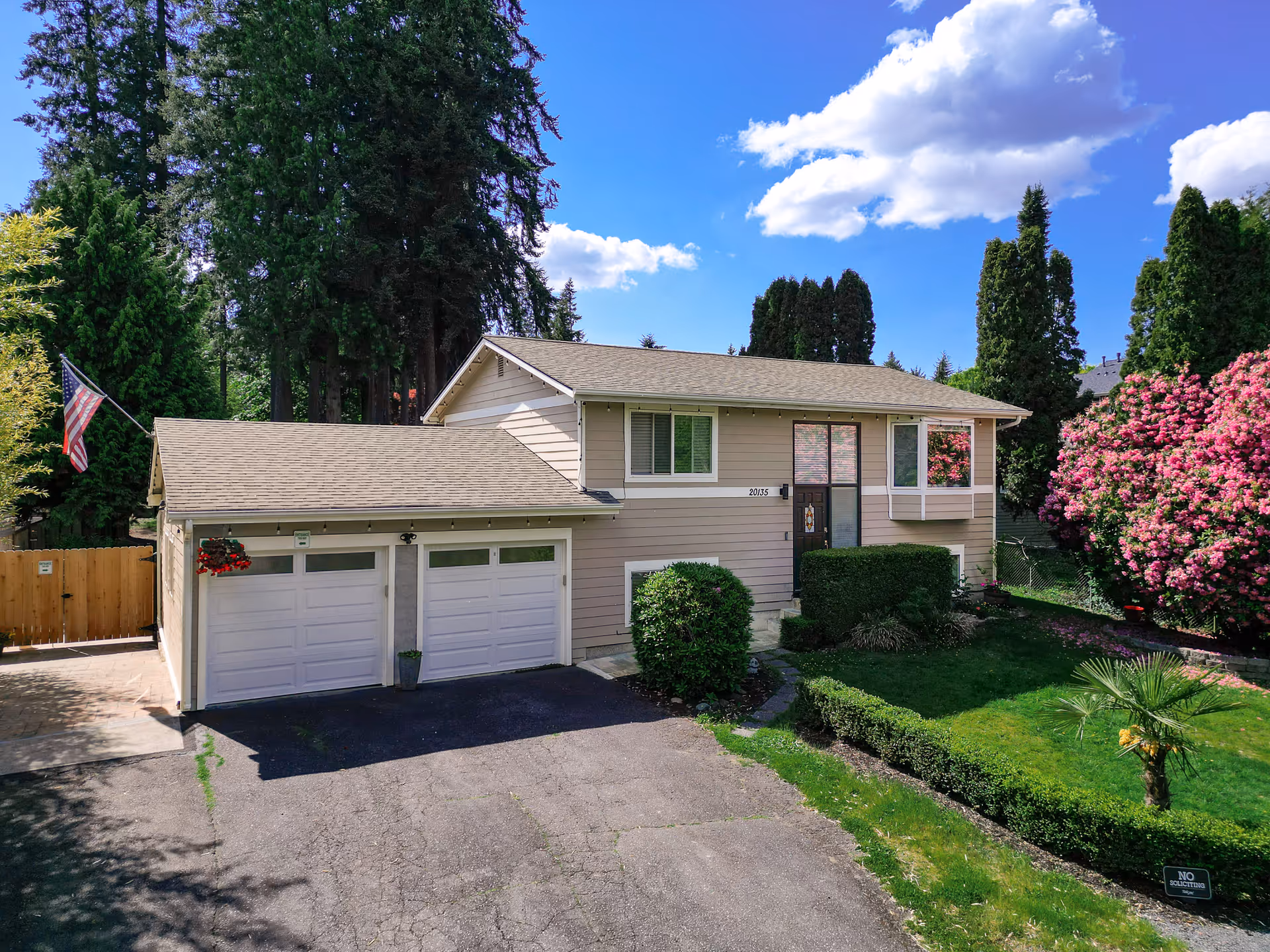 Two-story beige house with attached garage, manicured lawn, and flowering shrubs.