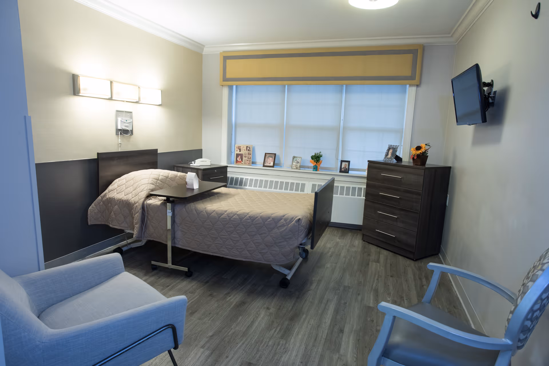 Hospital-style patient room with a bed, dresser, window, mounted TV, and seating.
