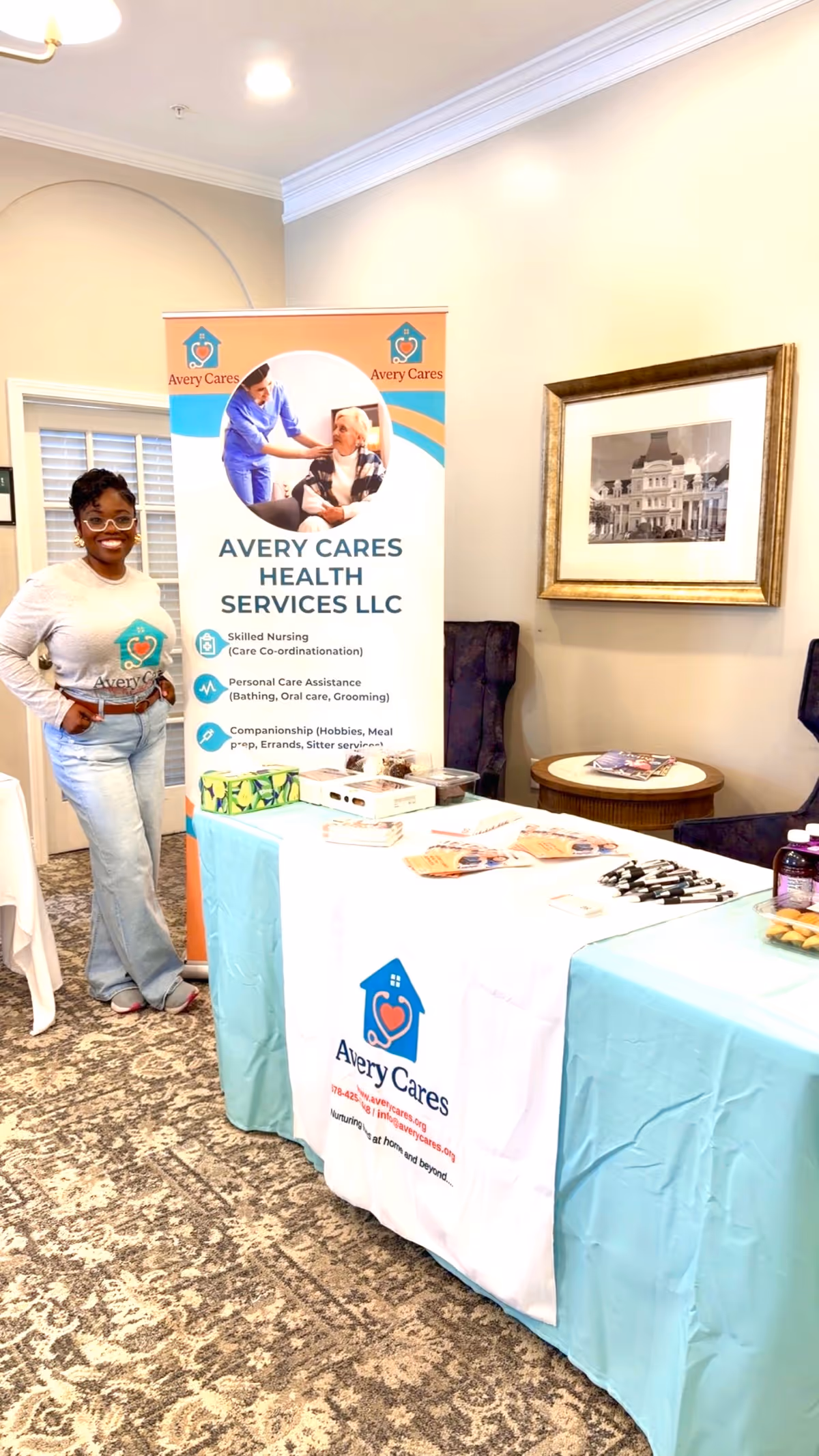 A smiling woman stands beside an Avery Cares Health Services banner at an indoor booth.