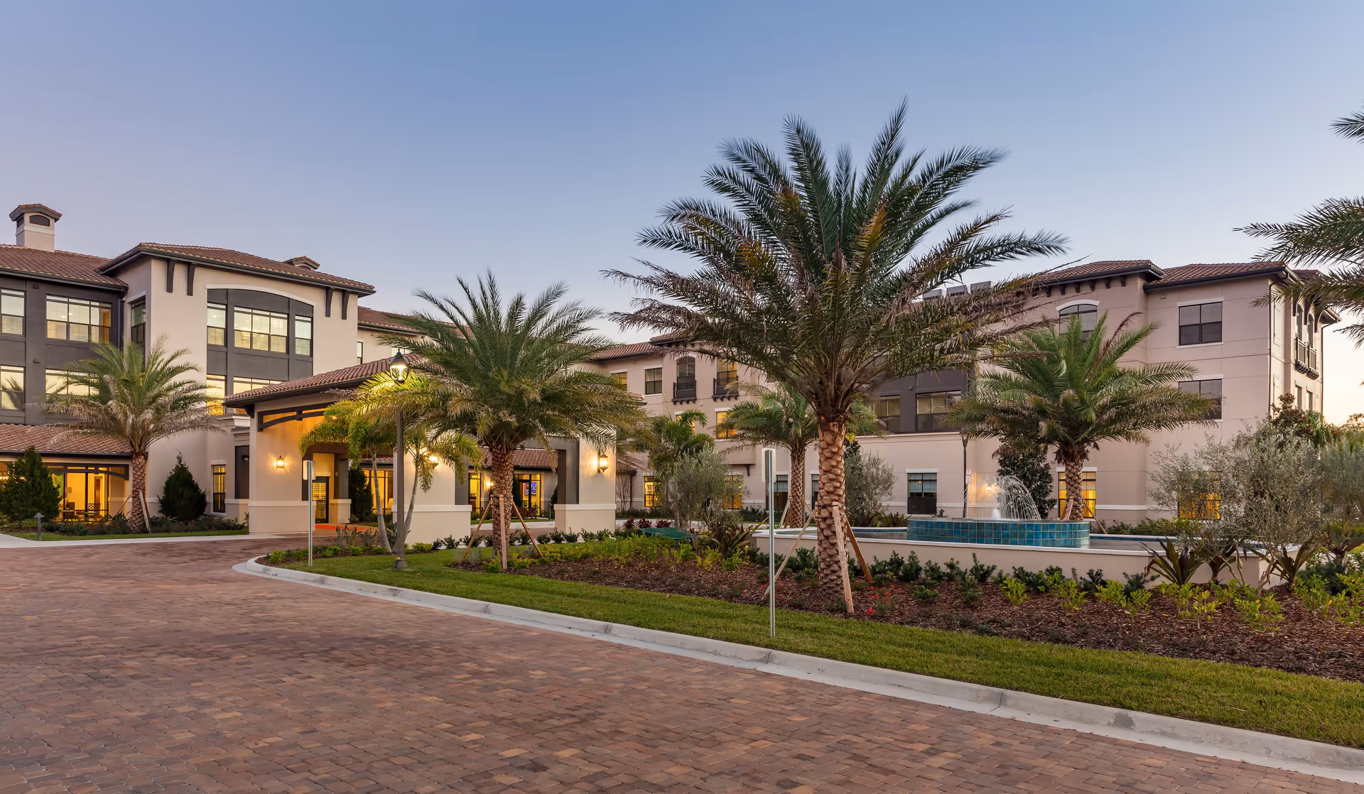 Exterior view of HarborChase of Dr. Phillips senior living facility at dusk, showing a multi-story building with large windows, palm trees, landscaped garden, and a fountain in front.