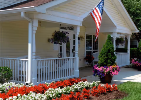 Front view of a porch with white railing, flower pots, and American flag at Arden Courts.