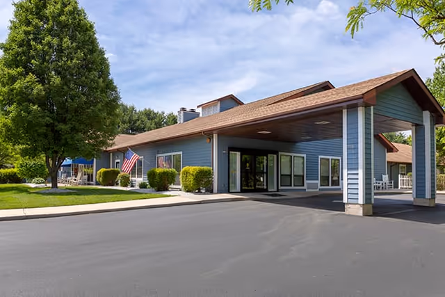 Exterior view of a single-story building with blue siding and a brown roof, featuring a covered entrance supported by columns. There is a large tree and neatly trimmed bushes near the building, an American flag displayed on the side, and a paved driveway in front under a partly cloudy sky.