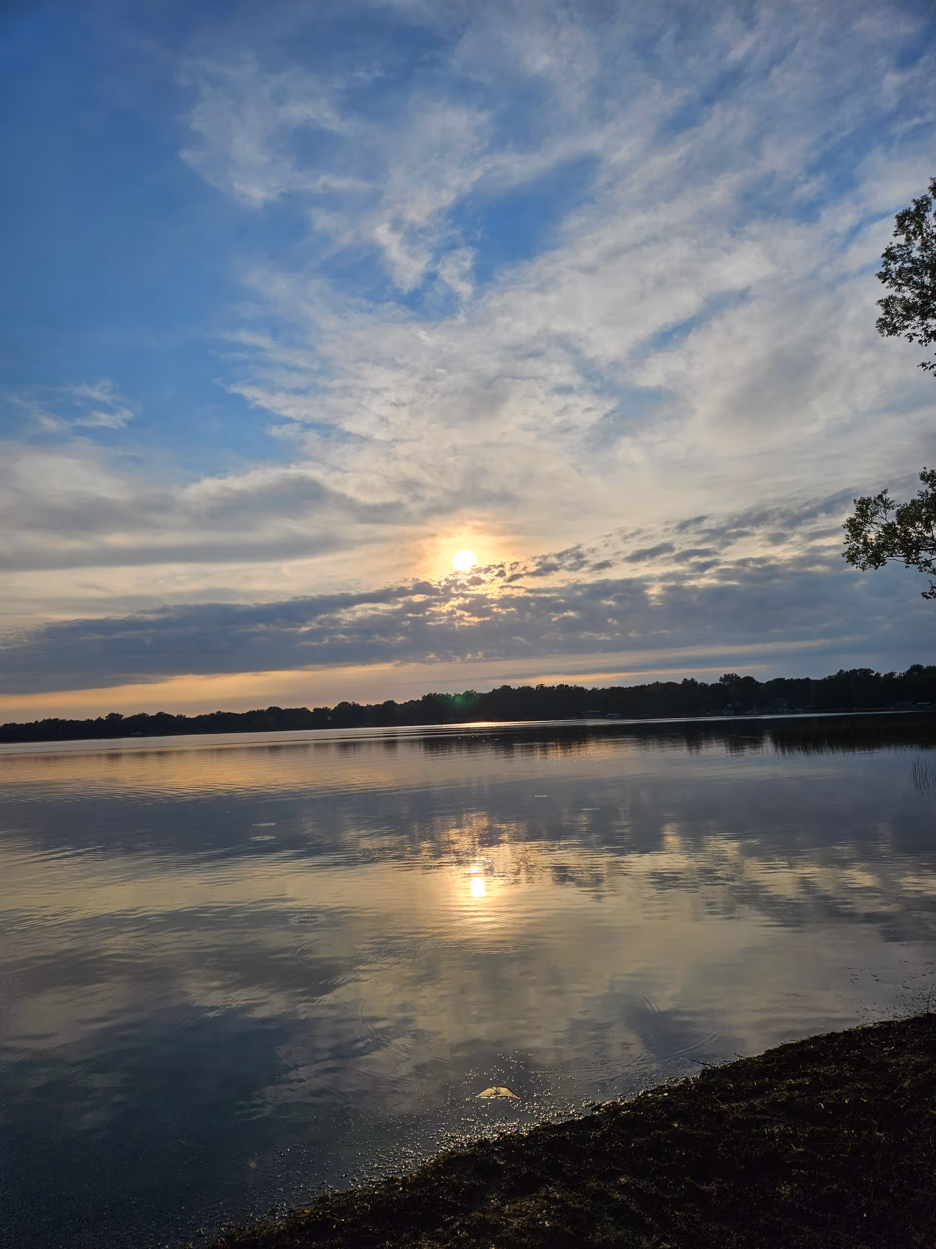 Sunset over a calm lake reflecting clouds with a distant tree line.