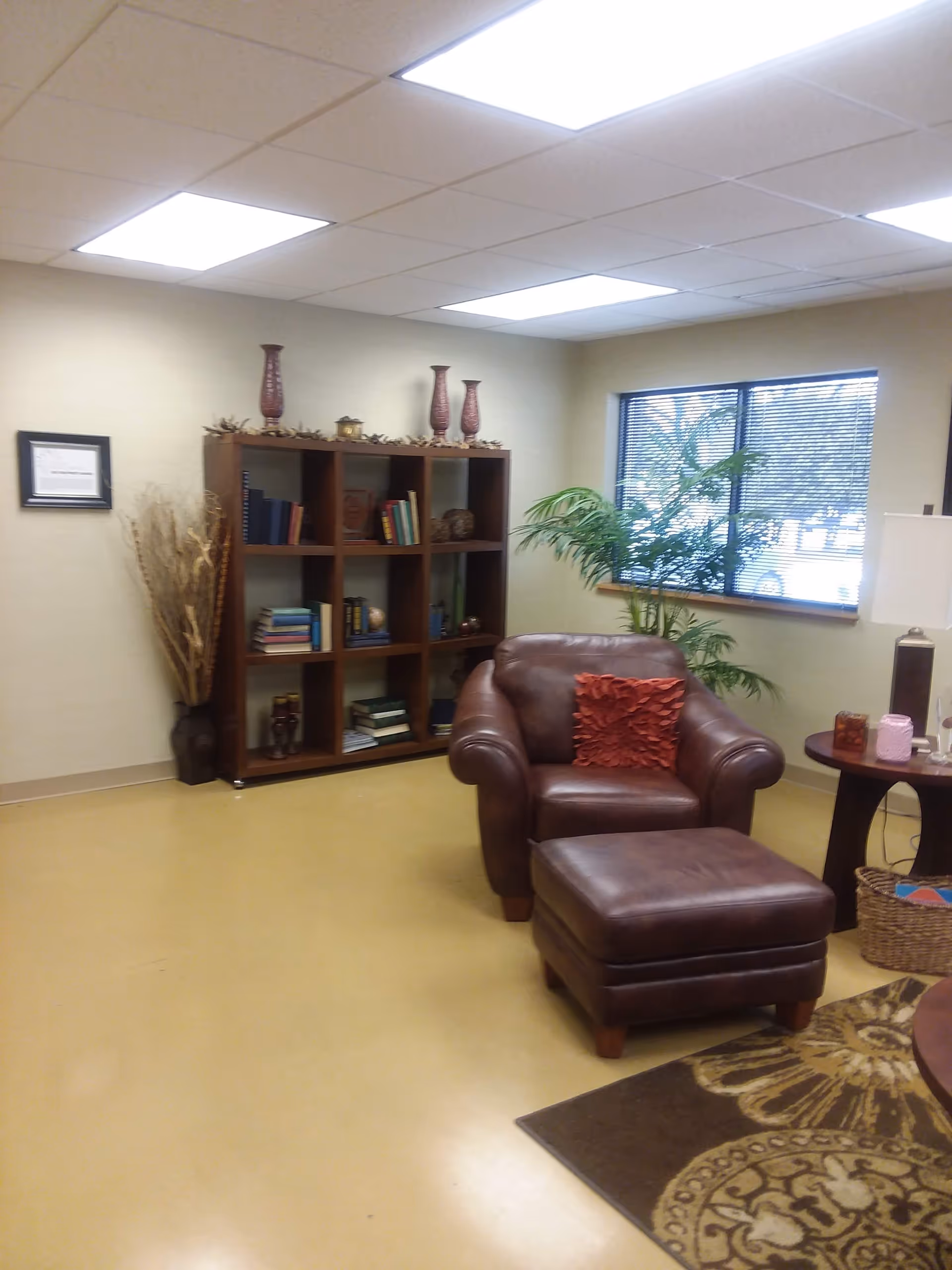 Cozy interior living room with leather armchair and ottoman, bookshelf, plant, and window blinds.
