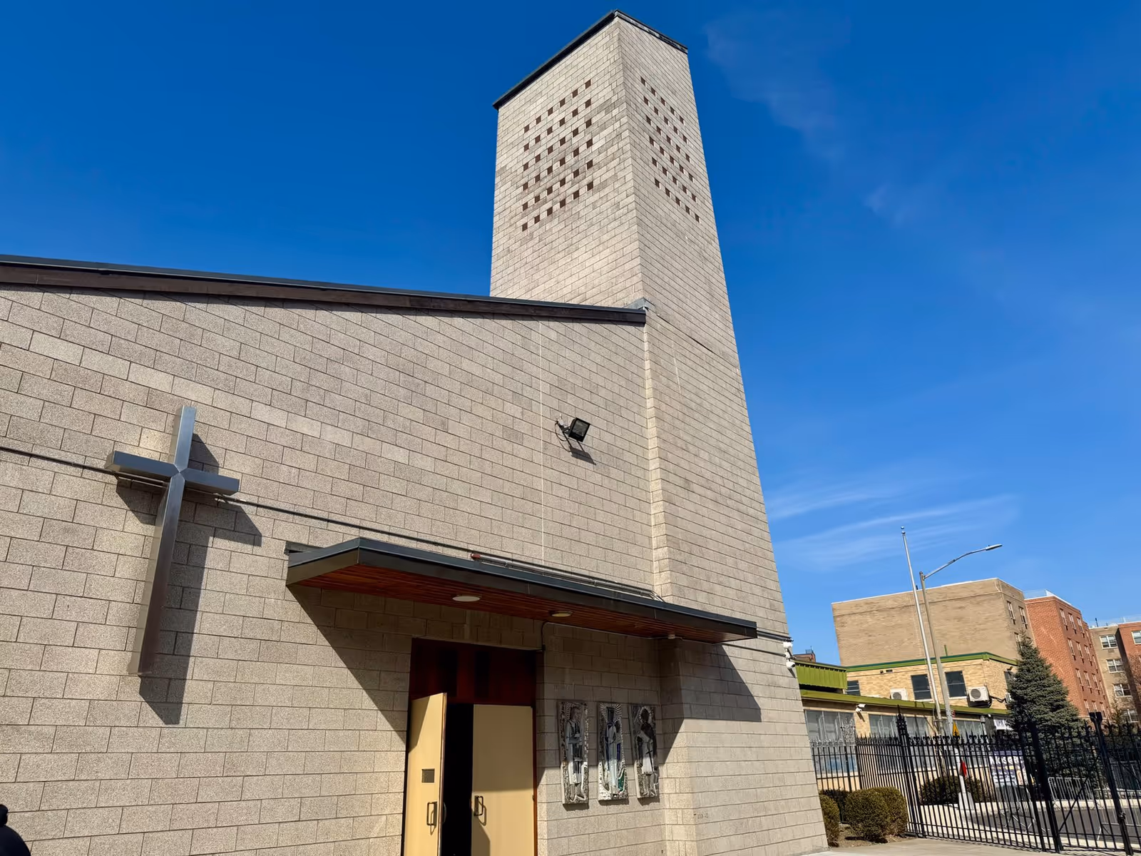 Exterior view of a beige brick church with a cross sculpture and entrance canopy.