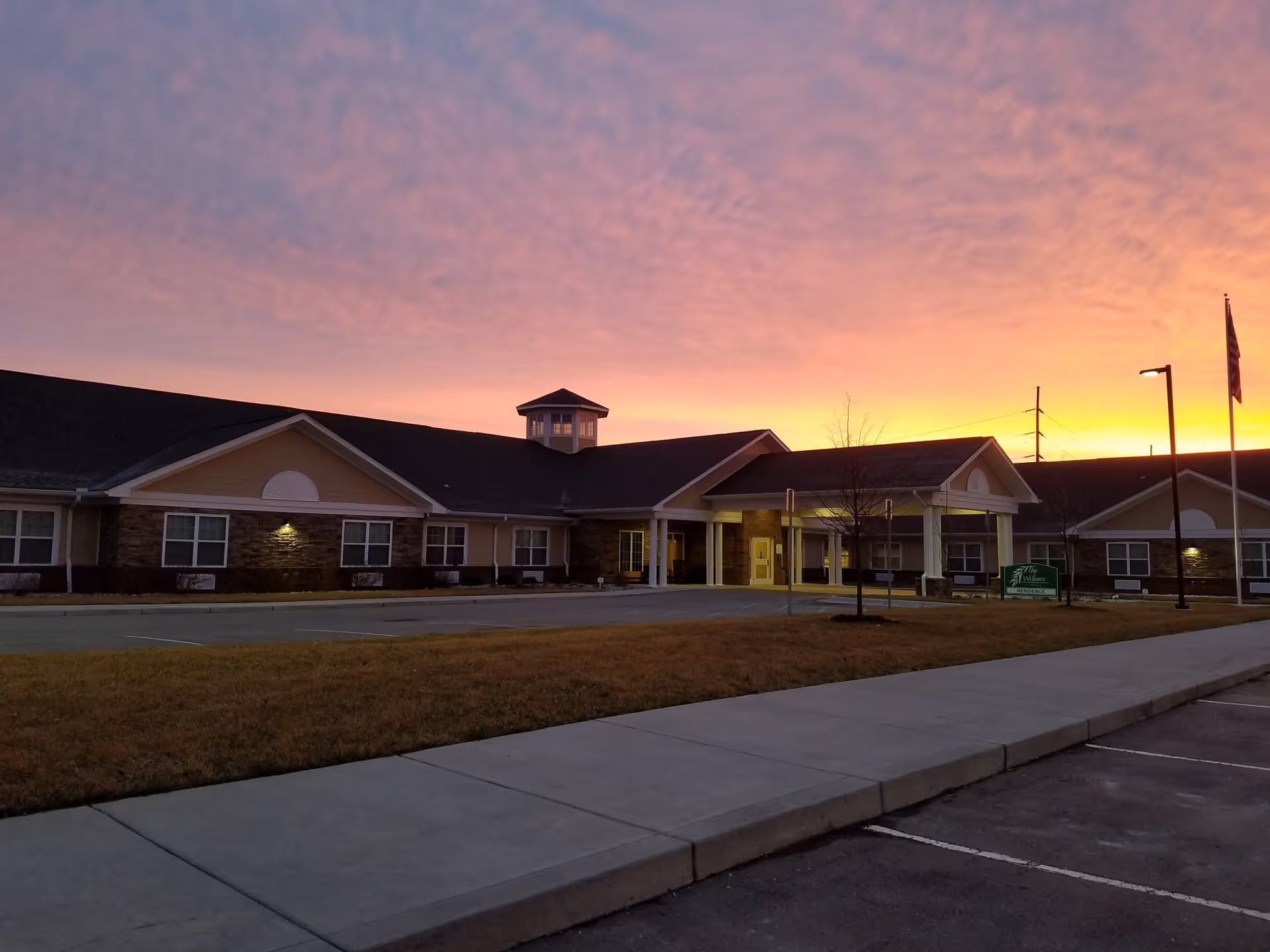 Exterior view of The Willows At Howell senior living facility at sunset, showing a single-story building with a covered entrance, windows, a flagpole with an American flag, and a parking area in front.