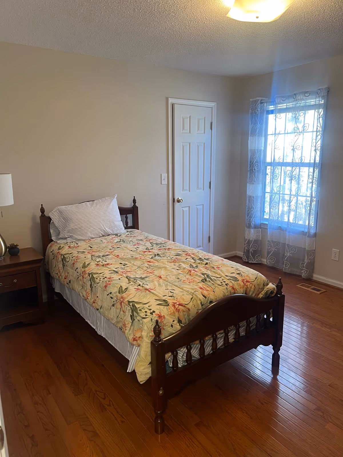 A single bed with a floral quilt, nightstand, lamp, and lace-curtained window in a tidy bedroom.